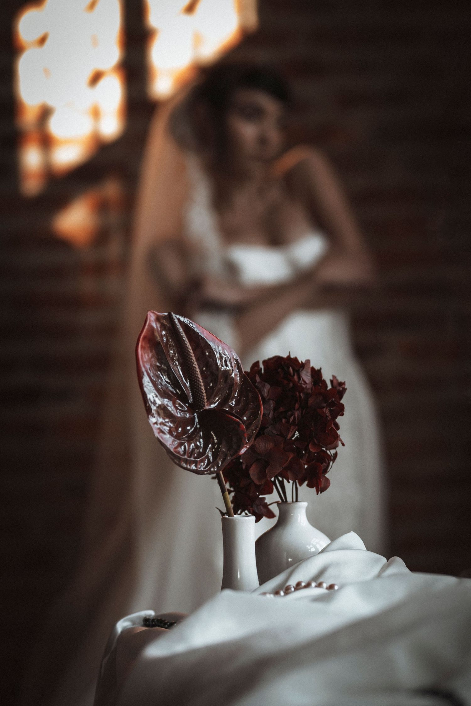 Dark red flowers in white vase with blurred figure in white dress in background during sunset.