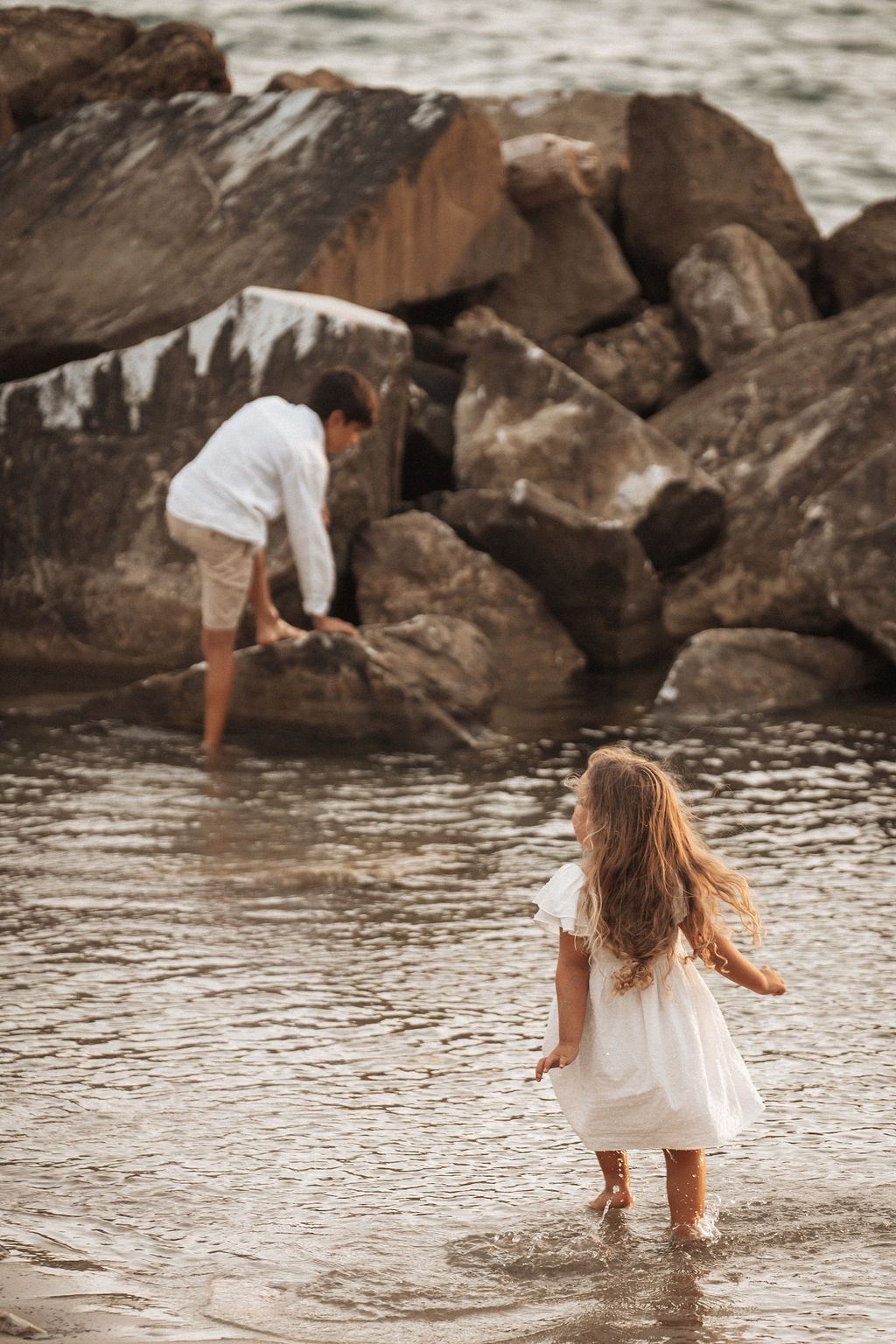 Two people playing in shallow water near rocky coastal formations at sunset.
