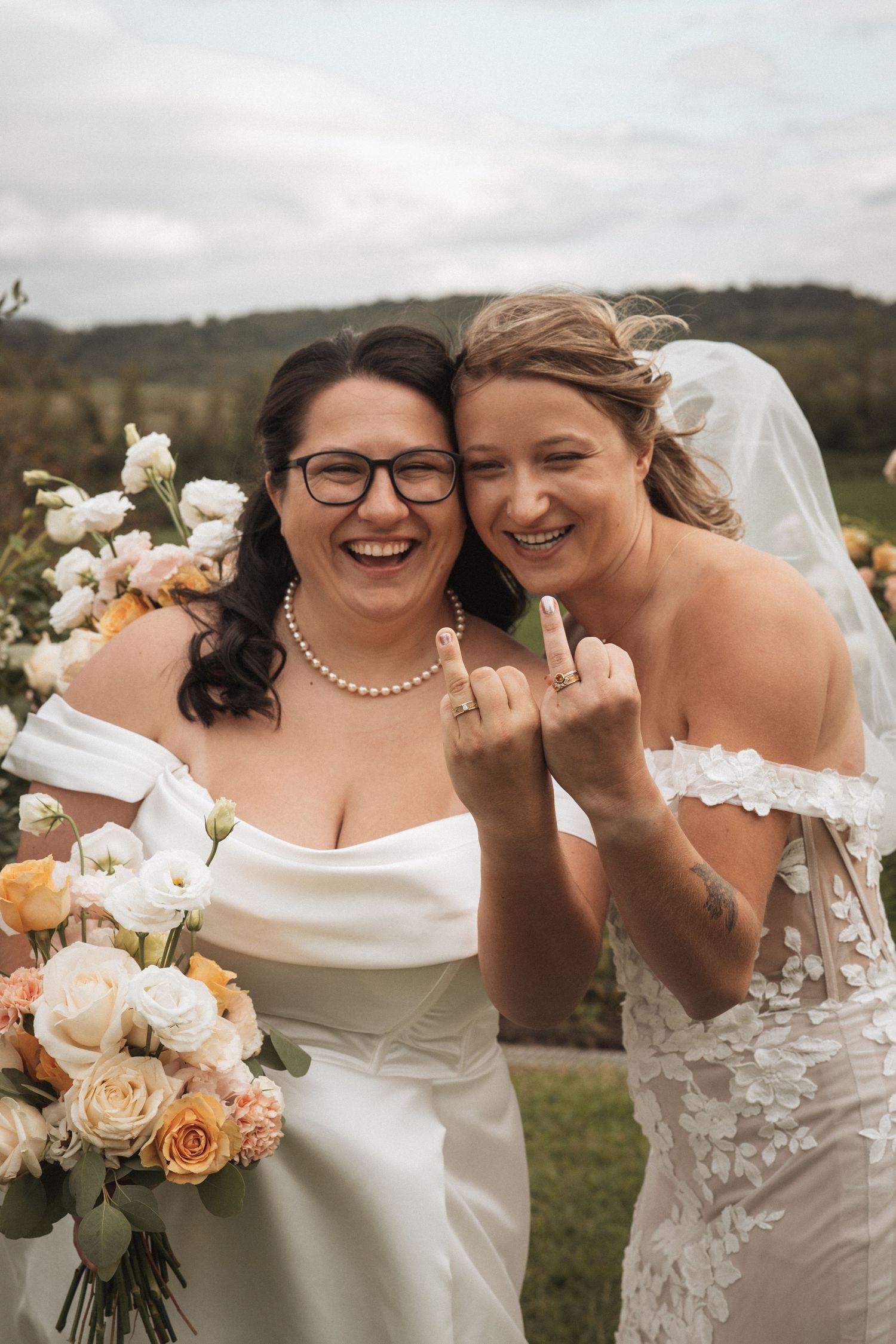 Two brides in white dresses show off their rings during an outdoor wedding celebration.