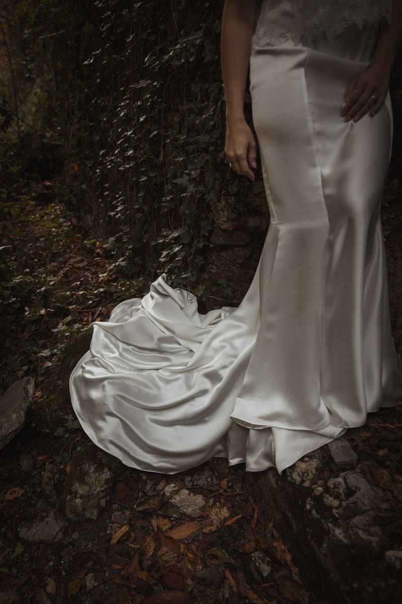 A flowing white wedding dress train trails elegantly across fallen autumn leaves in a forest setting.
