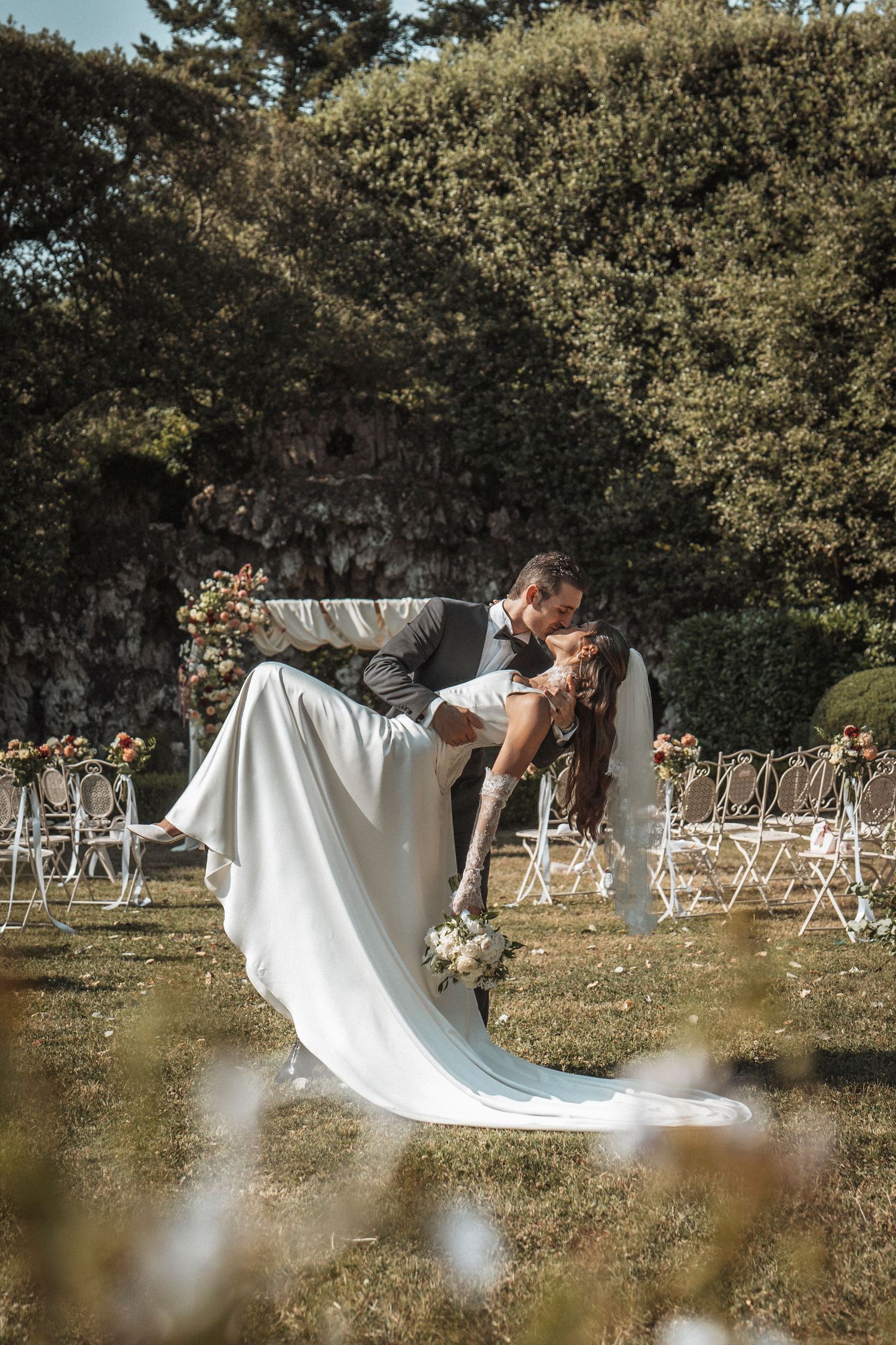 Groom dips bride in romantic outdoor wedding ceremony kiss, surrounded by floral arch and white chairs.
