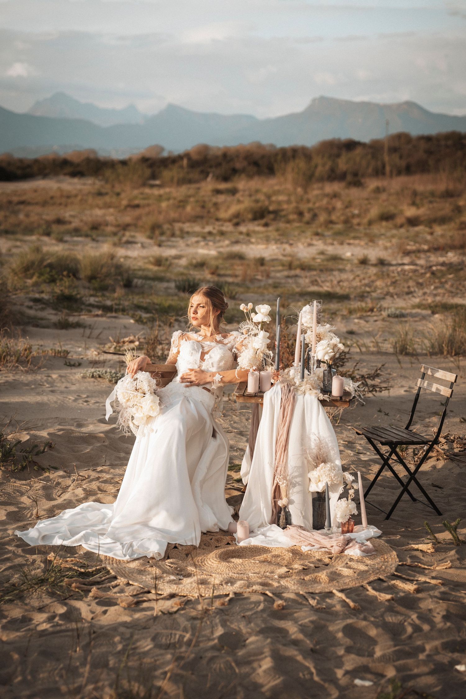 Romantic bridal scene in desert with white dress and vintage decor against mountain backdrop.