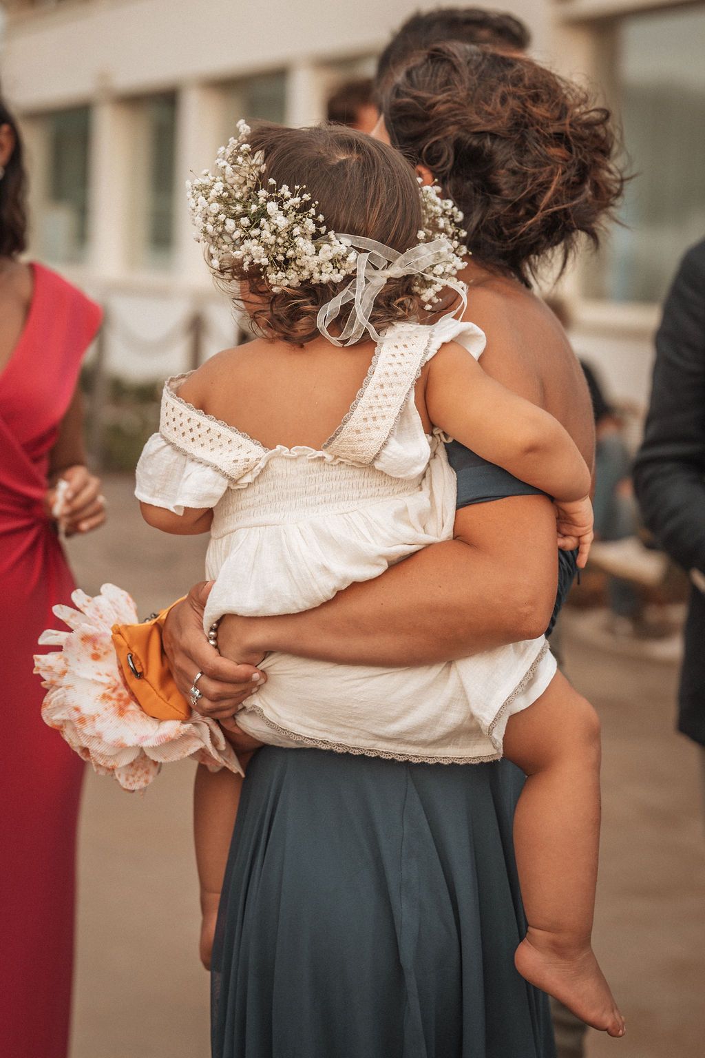 Person holding a small child wearing white dress and floral crown at a formal event.