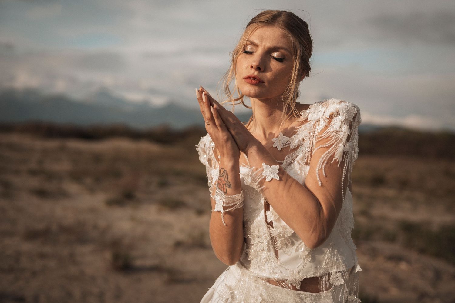 A person in a white lace wedding dress poses thoughtfully in a desert landscape at sunset.