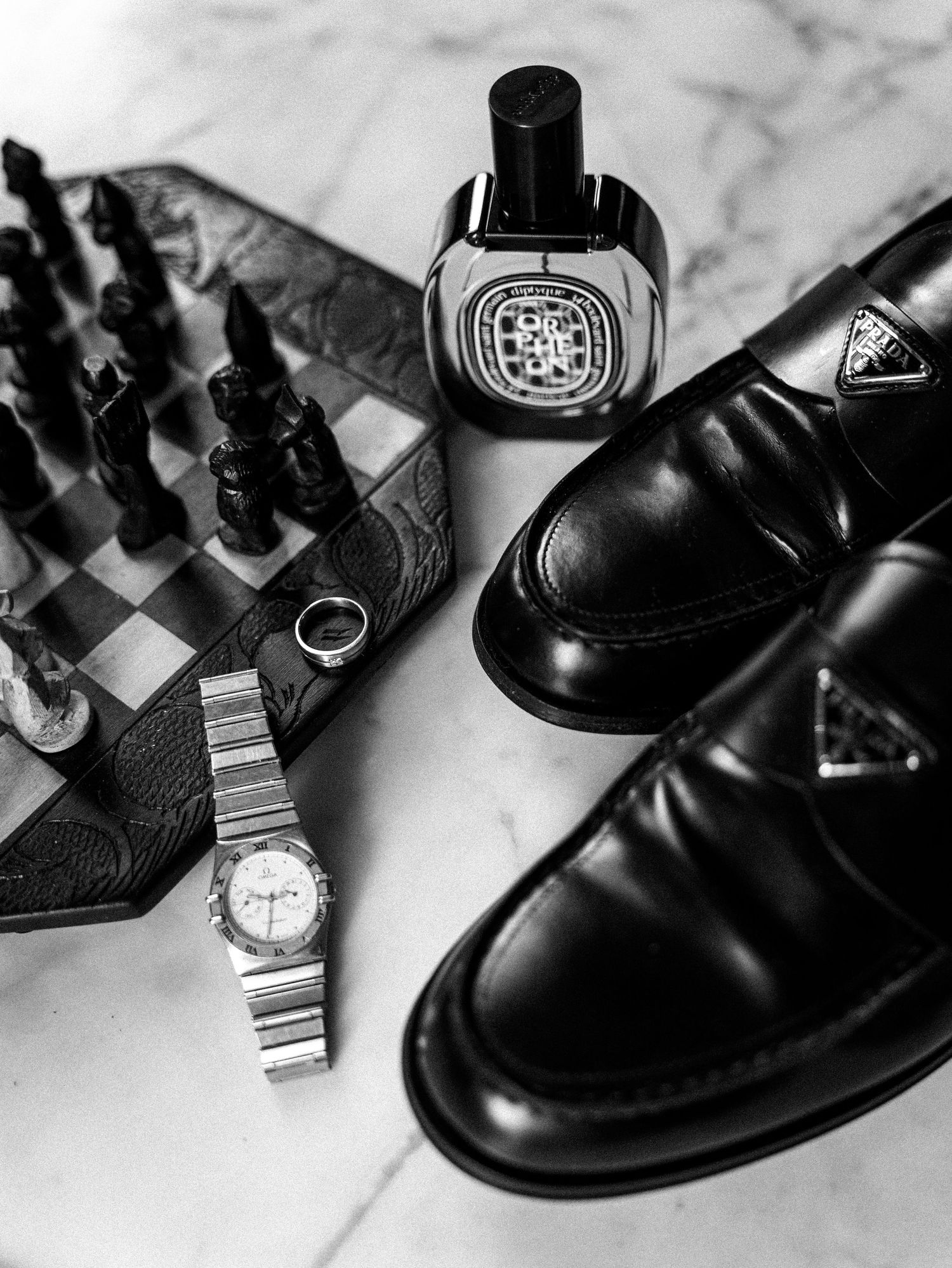 Black and white flat lay of Prada loafers, Diptyque perfume, Omega watch, chess set, and ring on marble.