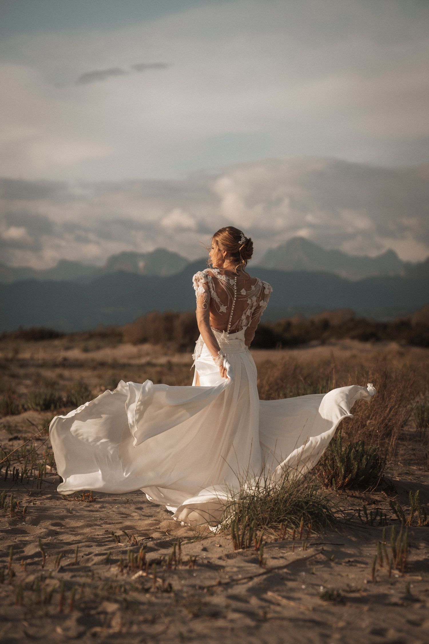 A flowing white wedding dress billows in the wind against a mountainous landscape at sunset.