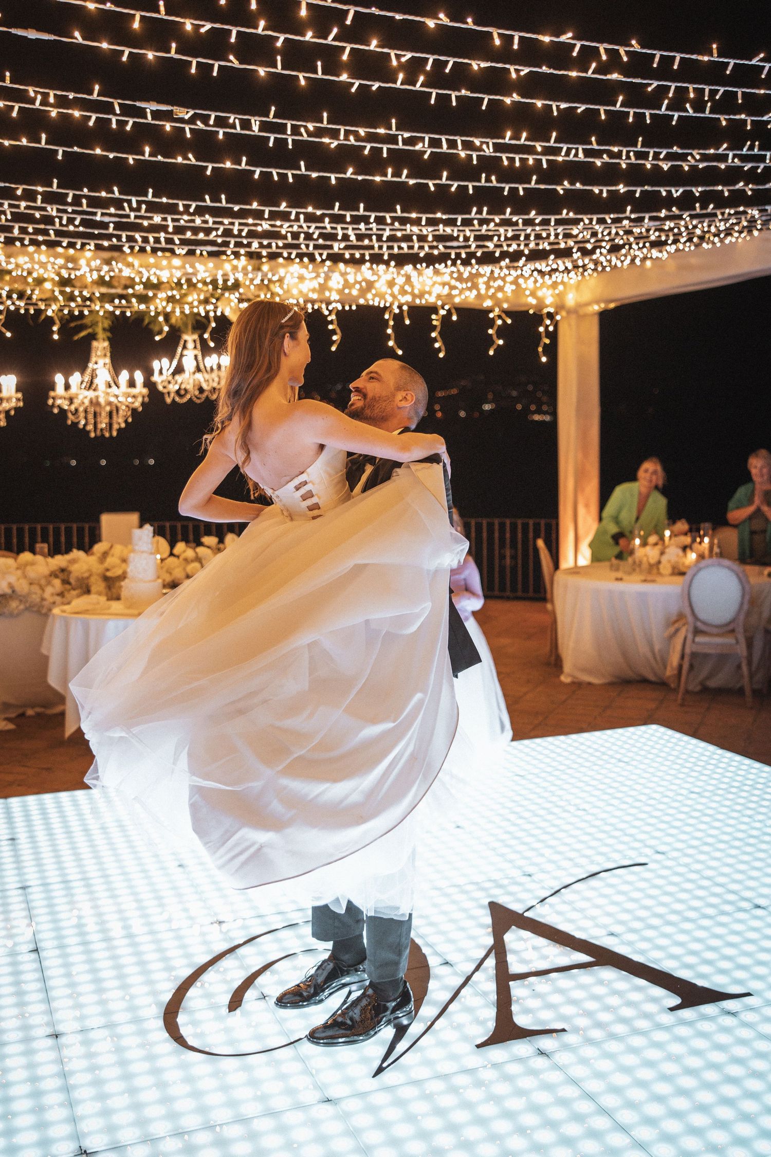 Couple dances under twinkling string lights on an illuminated dance floor at night.