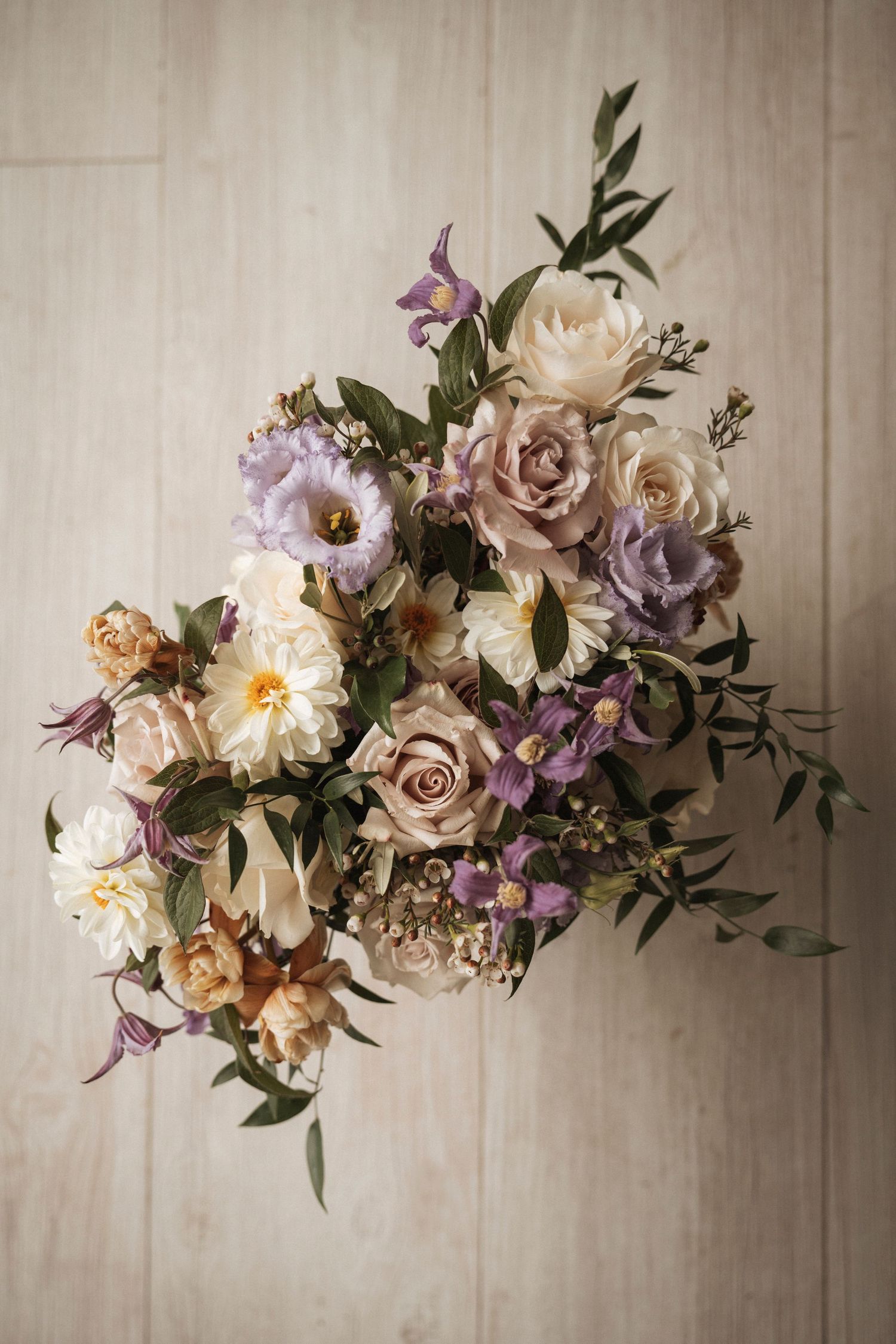Elegant floral arrangement featuring blush roses, white peonies and purple blooms against a neutral wall.