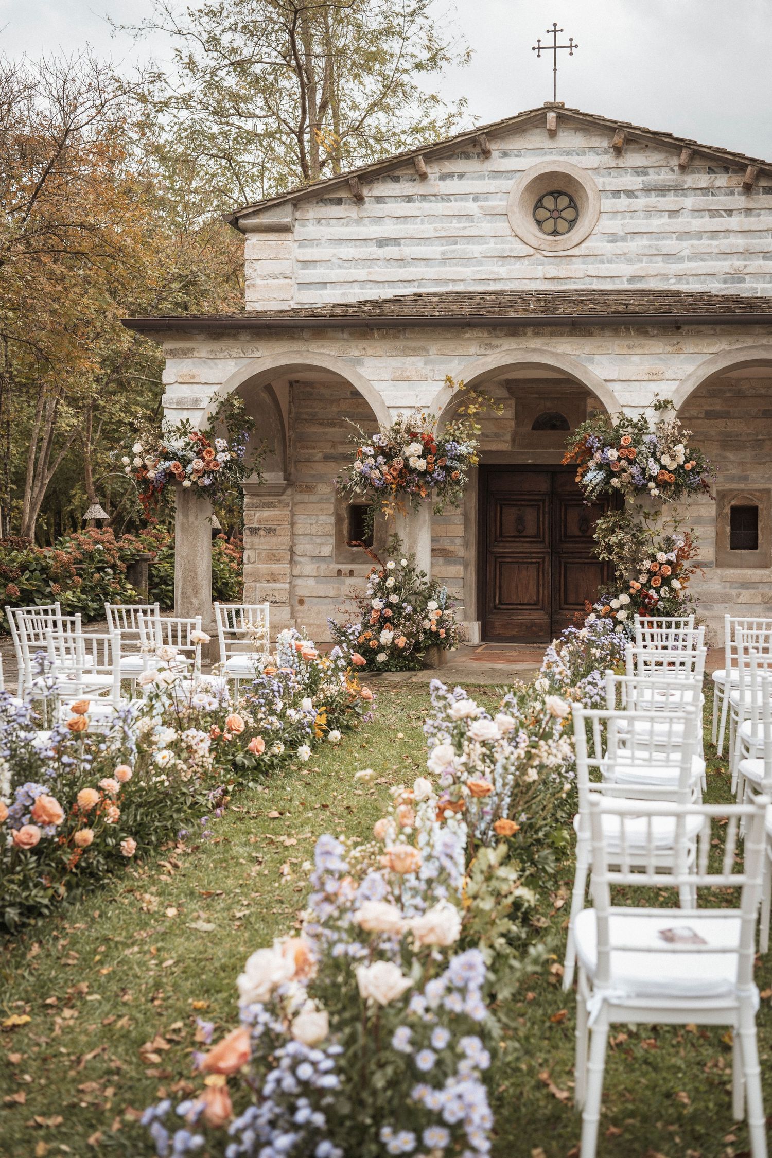 A rustic stone chapel with white wooden chairs and colorful floral arrangements set up for an outdoor wedding ceremony.