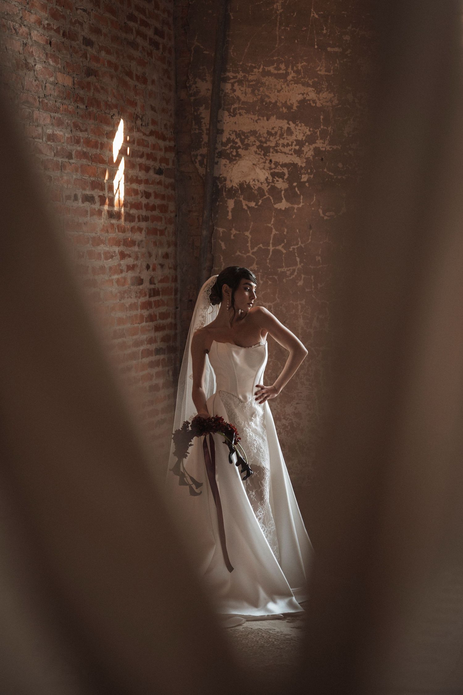 Artistic bridal portrait series showing flowing white dress against rustic brick wall with dramatic lighting.