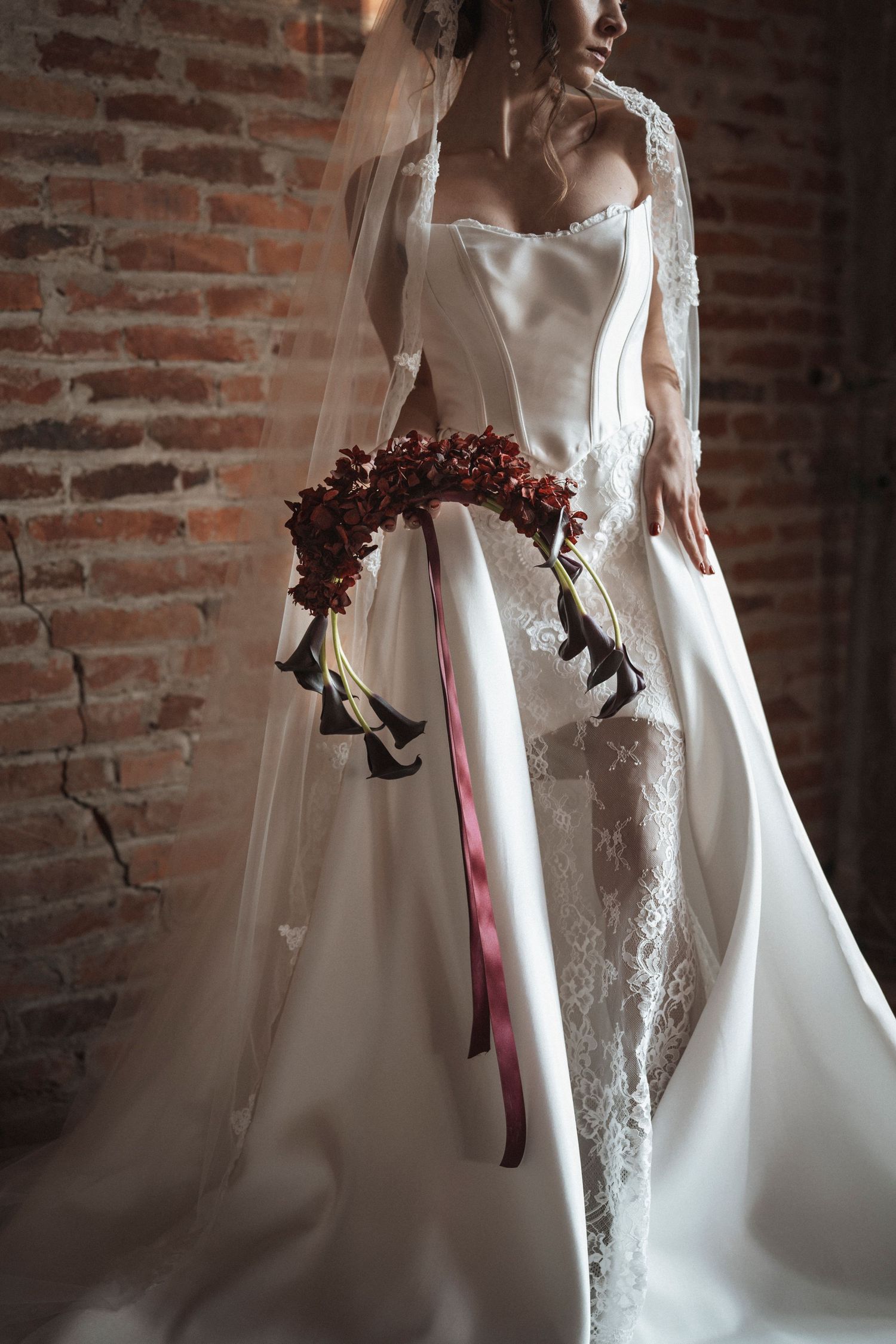 Elegant bride in white lace gown holding burgundy floral bouquet against rustic brick wall.