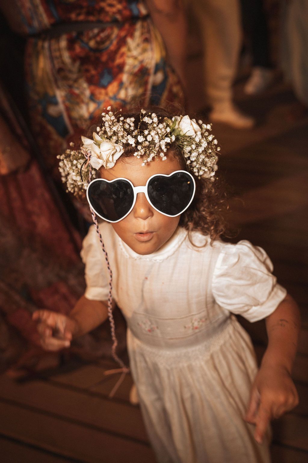 Child in white dress wearing heart-shaped sunglasses with flower crown.