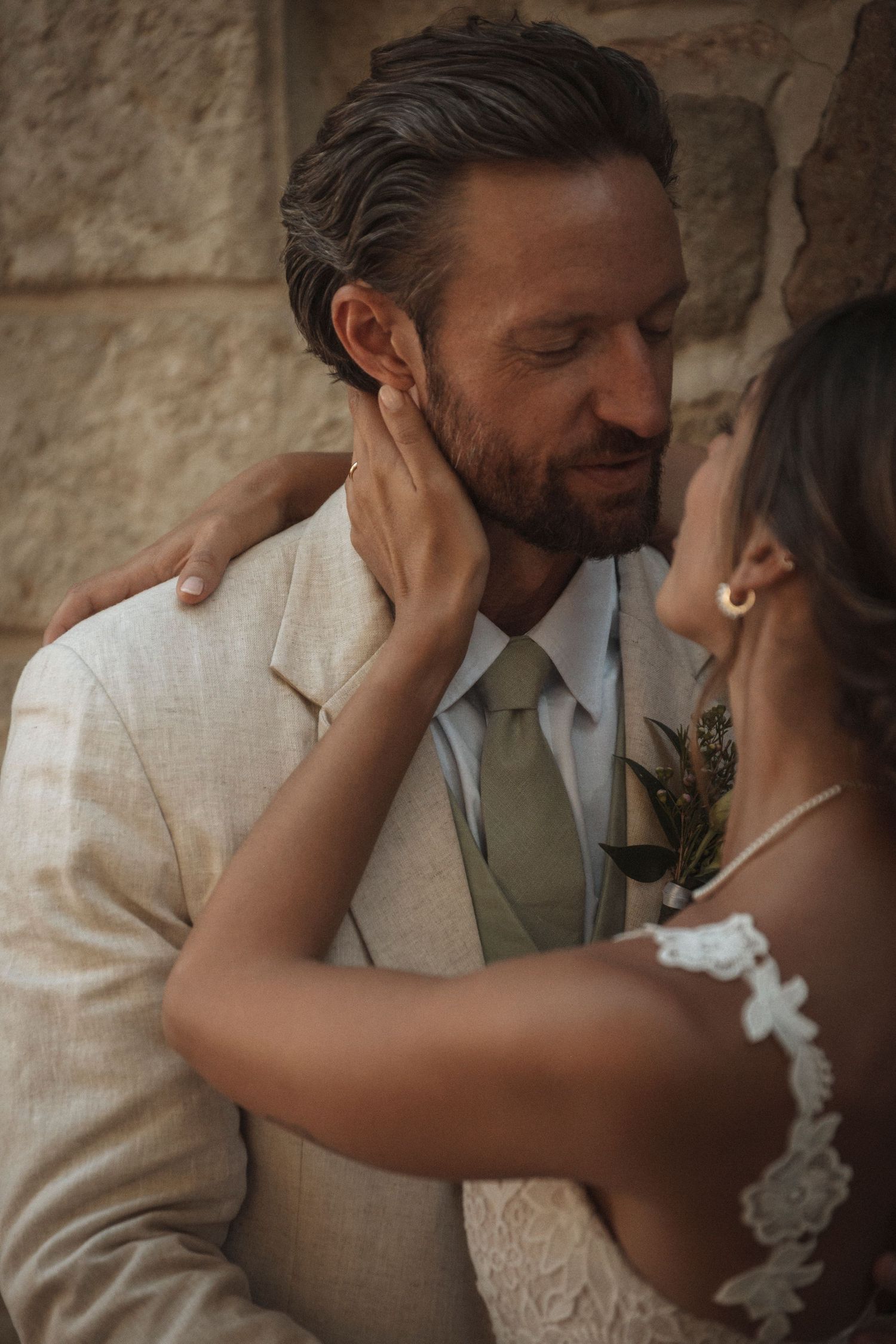 Intimate moment between couple in formal wedding attire against stone wall.