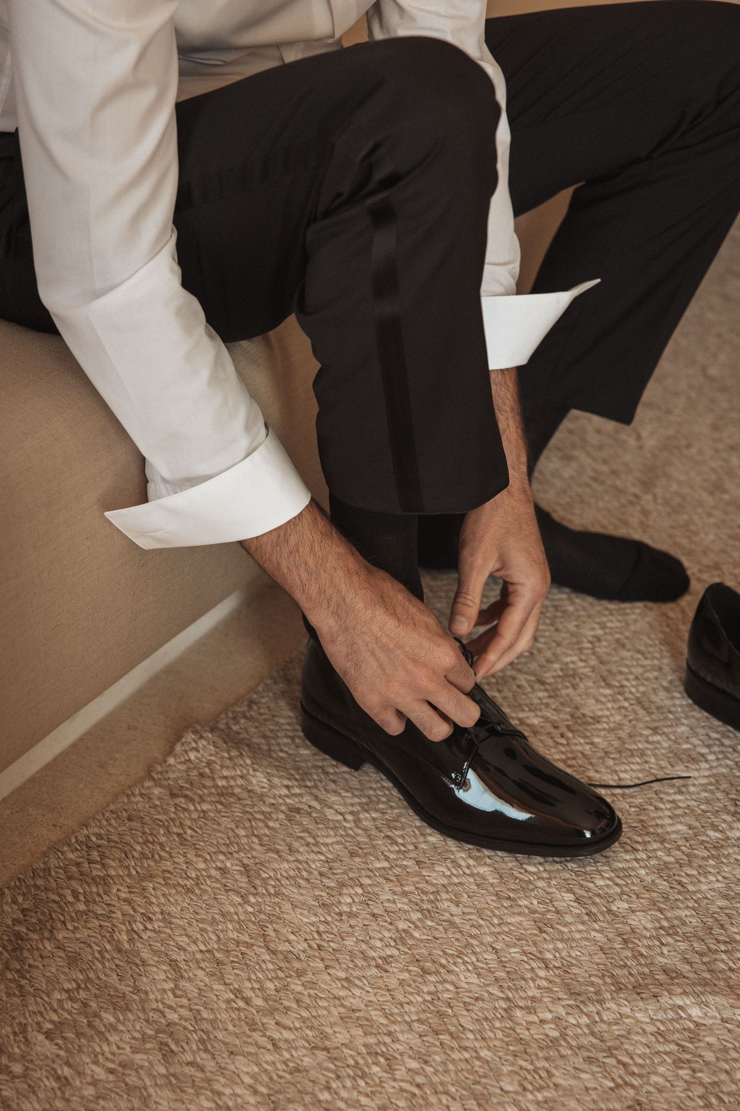 Person in formal attire putting on black dress shoes on carpeted floor.