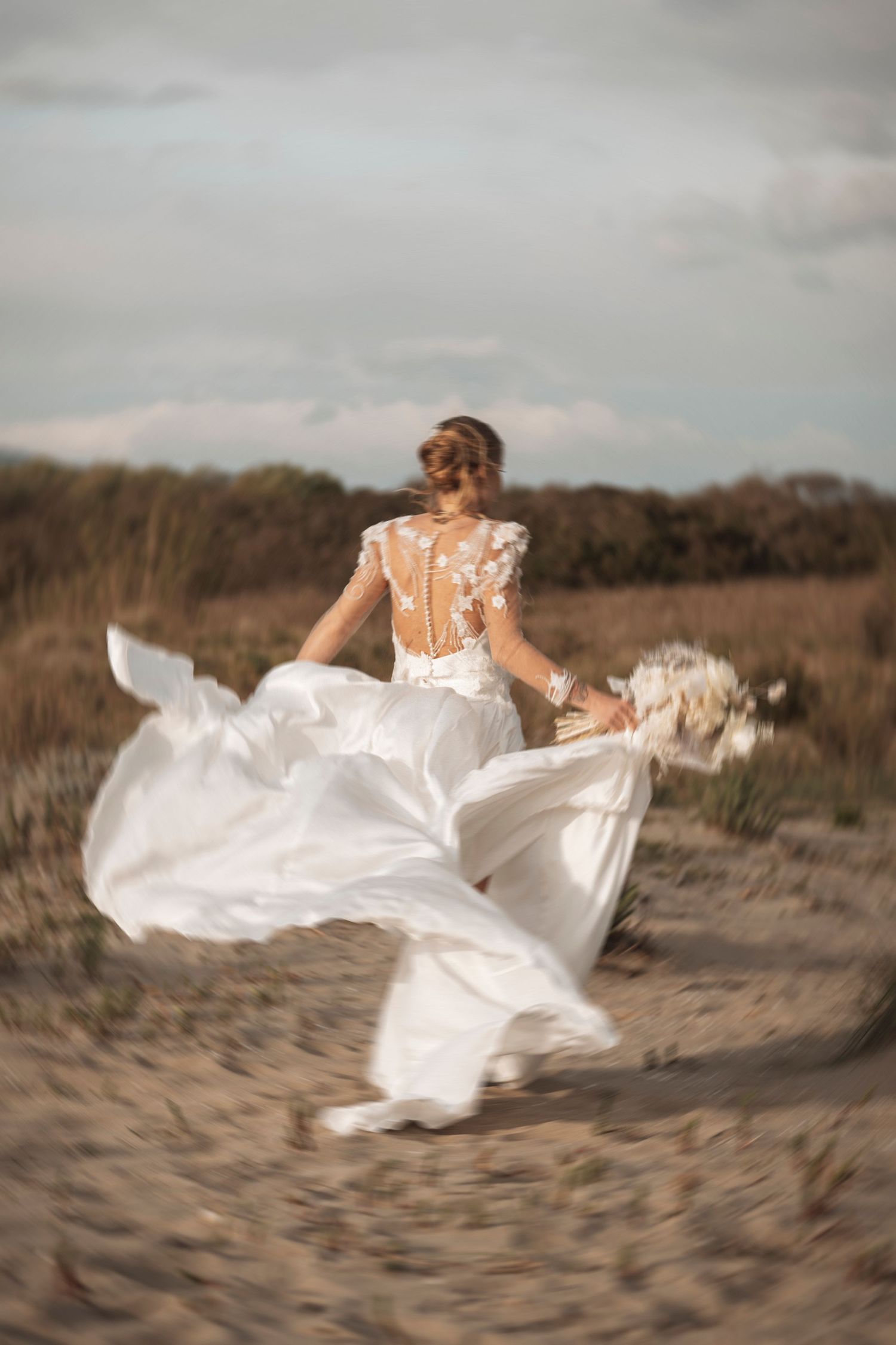 A bride's dress billows dramatically as she spins on a sandy beach with stormy clouds overhead.