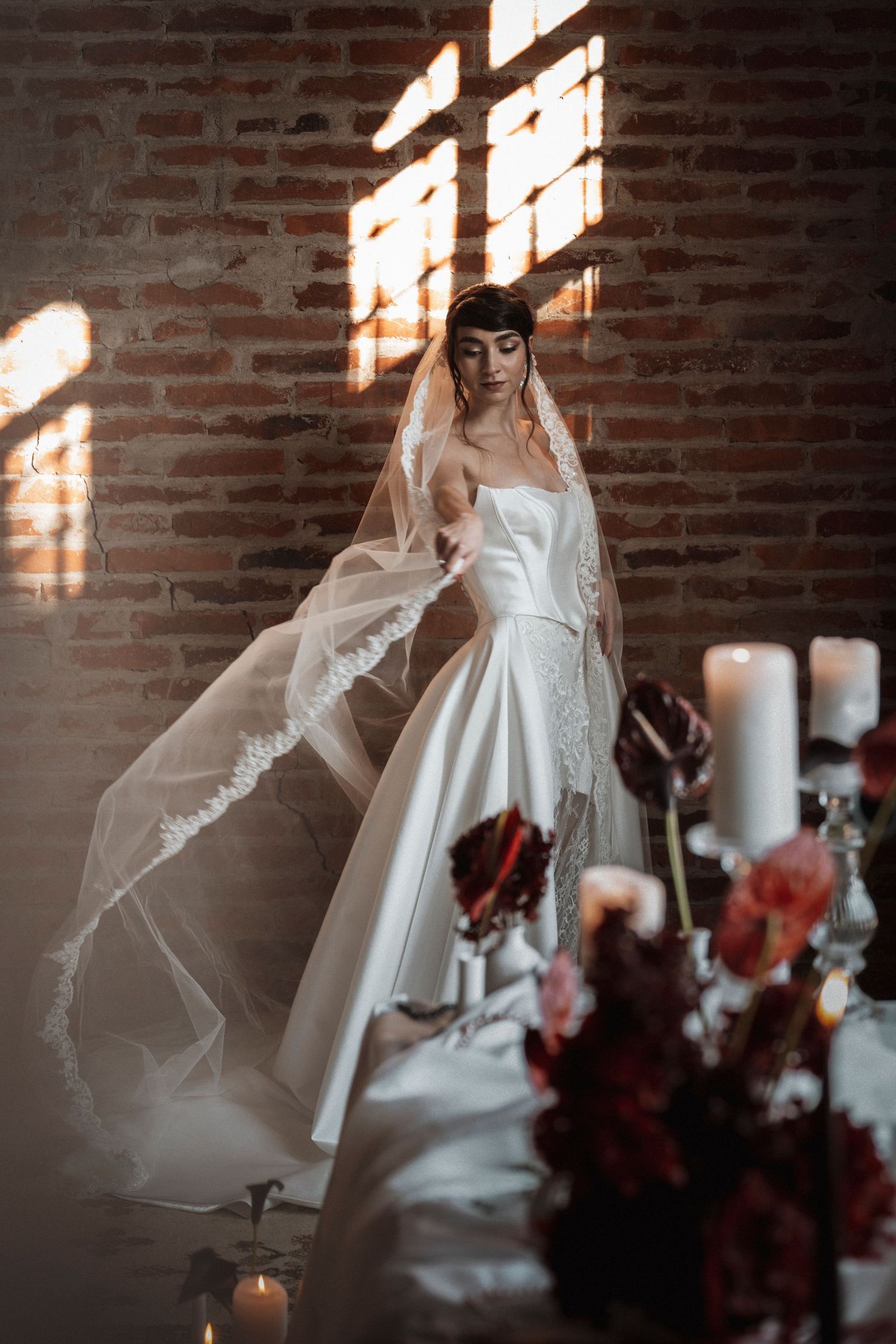 A bride in a white wedding dress stands against a brick wall with dramatic window light casting shadows.