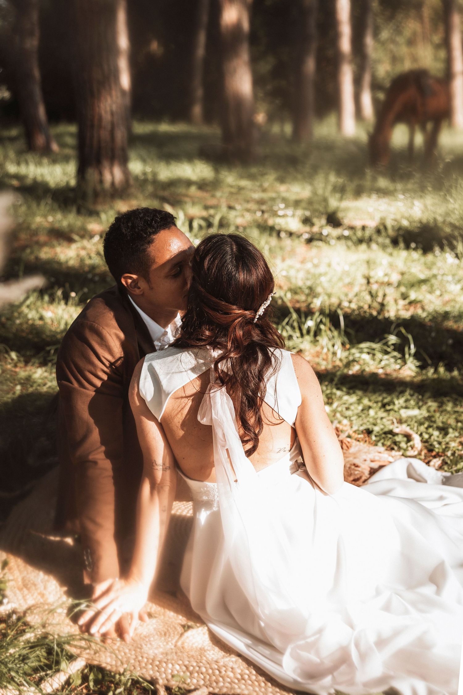 Couple in wedding attire laying in sunlit forest clearing with dappled light through trees.