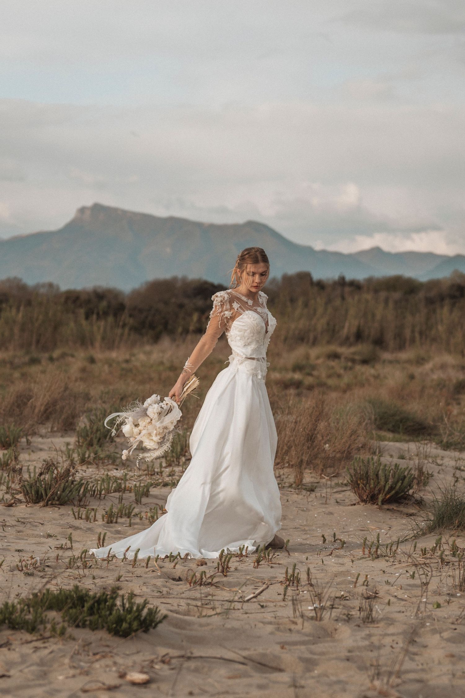 A flowing white wedding dress catches the wind against a mountainous desert landscape at sunset.