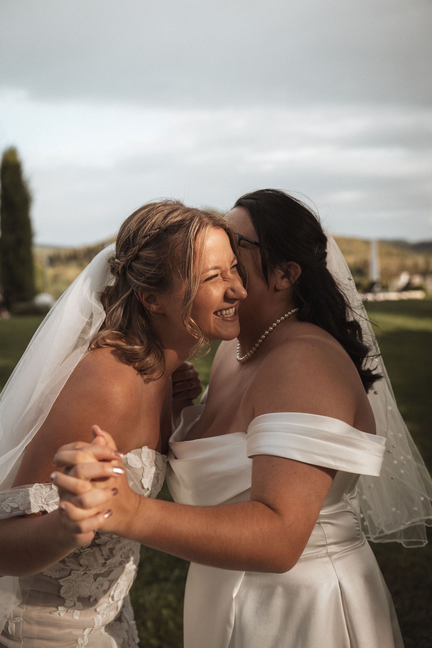 Two brides in white dresses share a joyful embrace outdoors under cloudy skies.