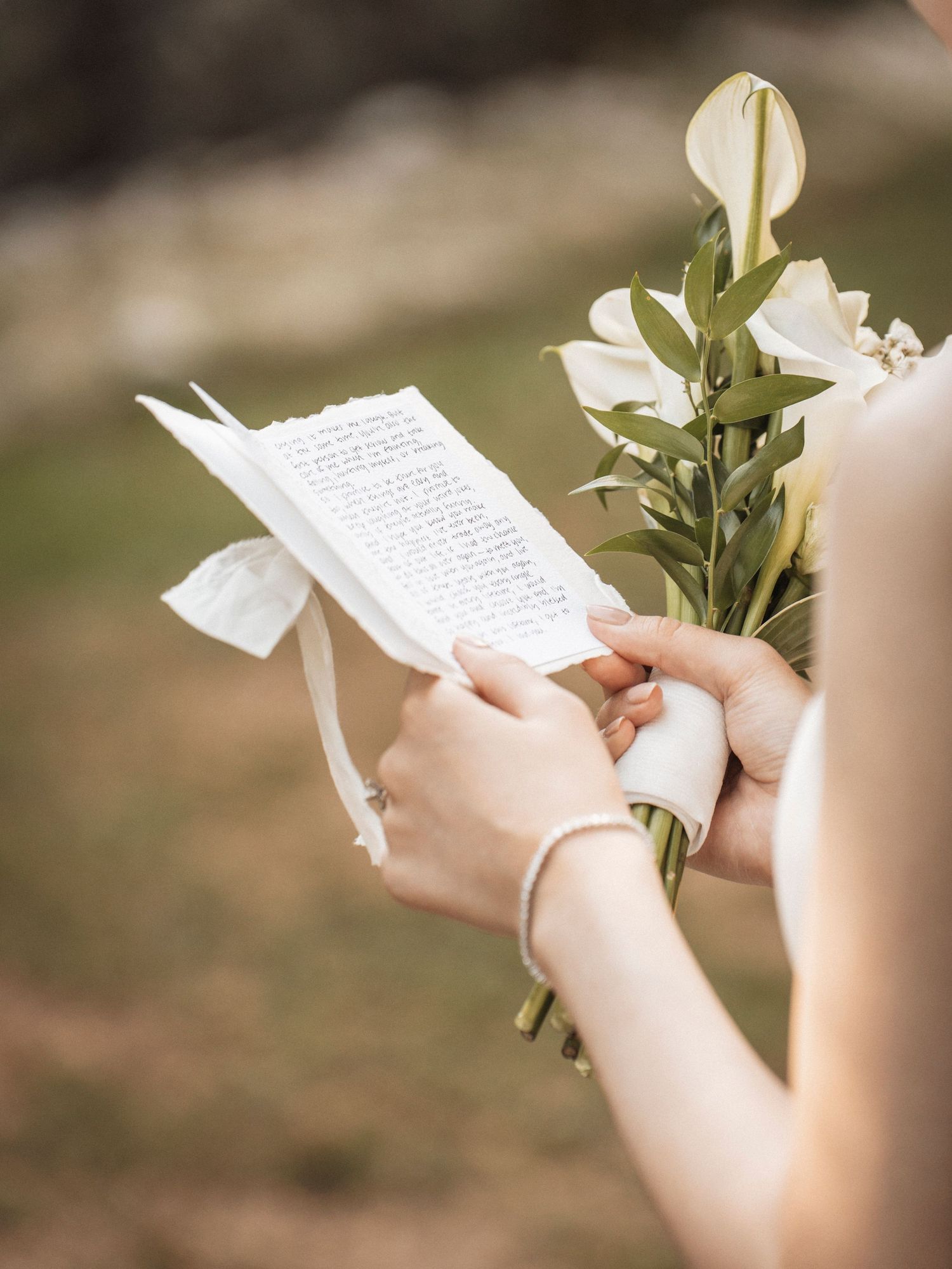 Bride holding white calla lily bouquet and reading vows card with ribbon at outdoor wedding ceremony.