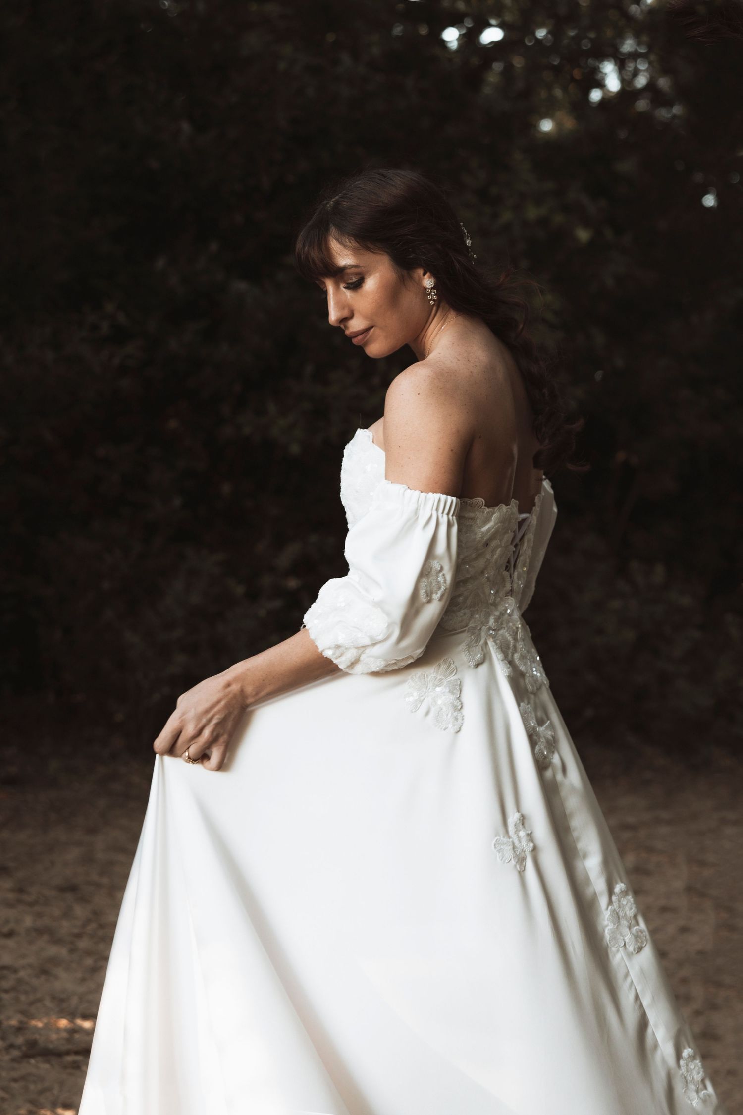 A bride in an elegant off-shoulder white wedding gown with lace details poses gracefully in a natural outdoor setting.