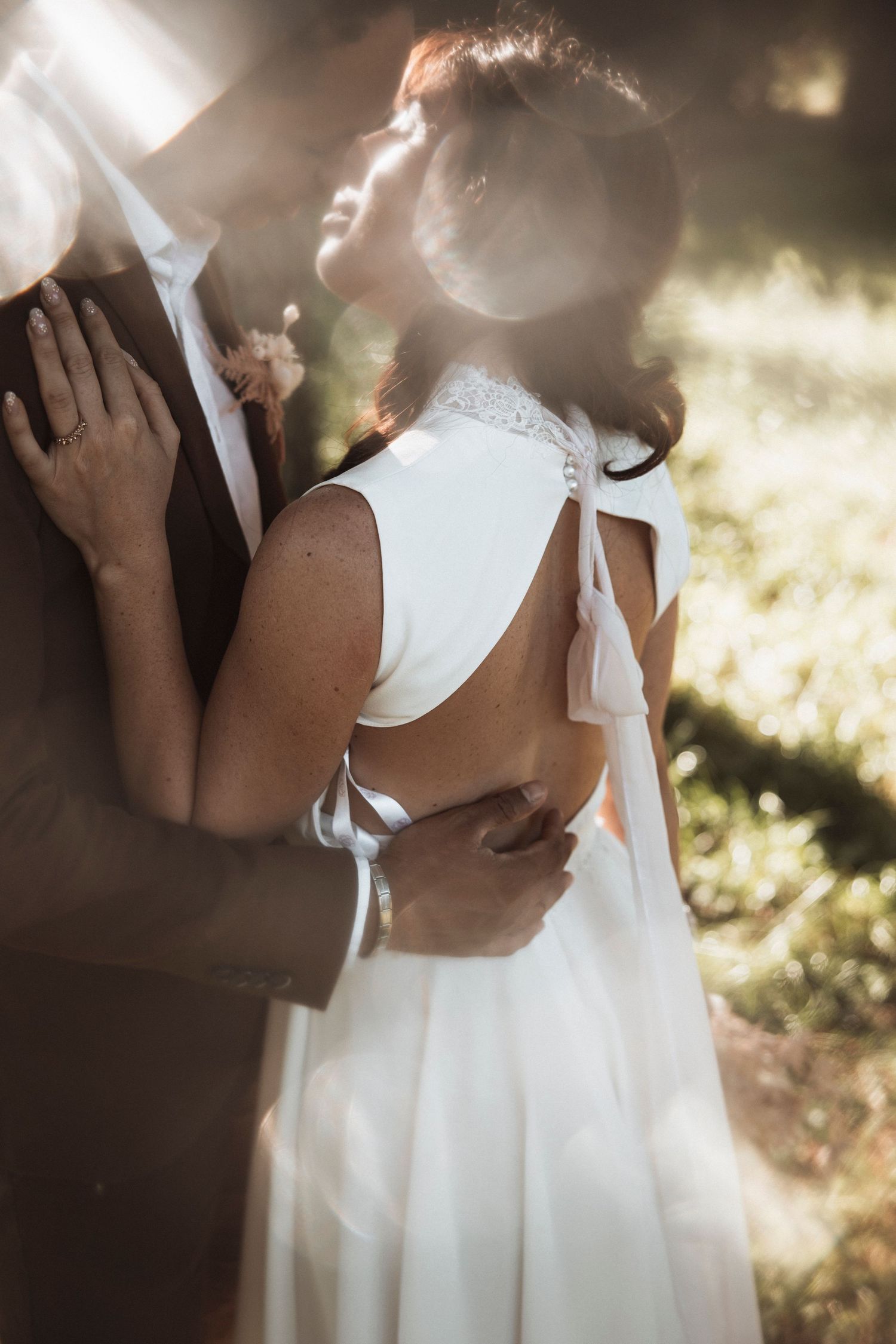Intimate wedding couple moment with white dress and dark suit in soft sunlit outdoor setting.