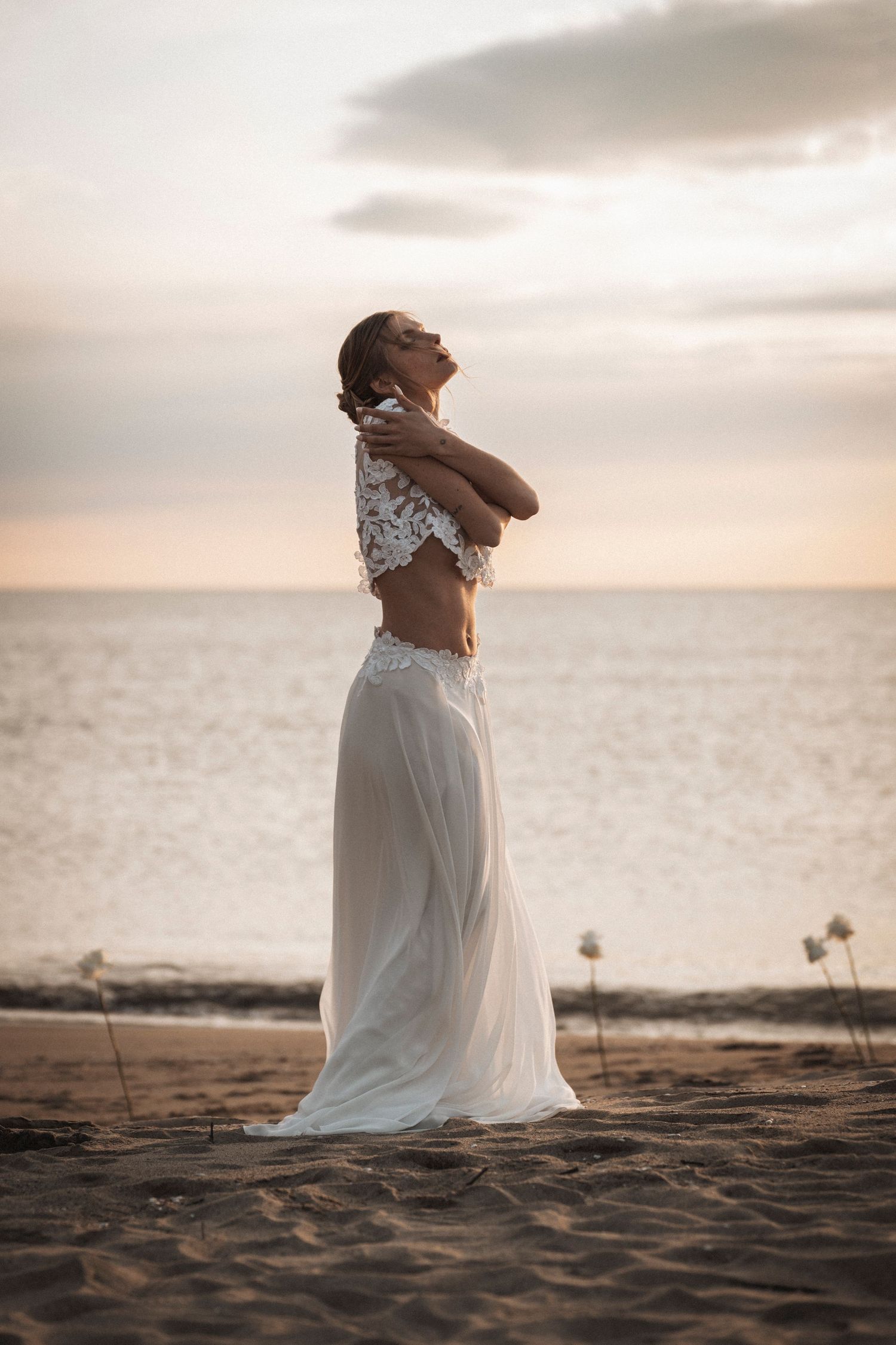A person in a flowing white dress stands on a beach at sunset with arms raised in a peaceful pose.