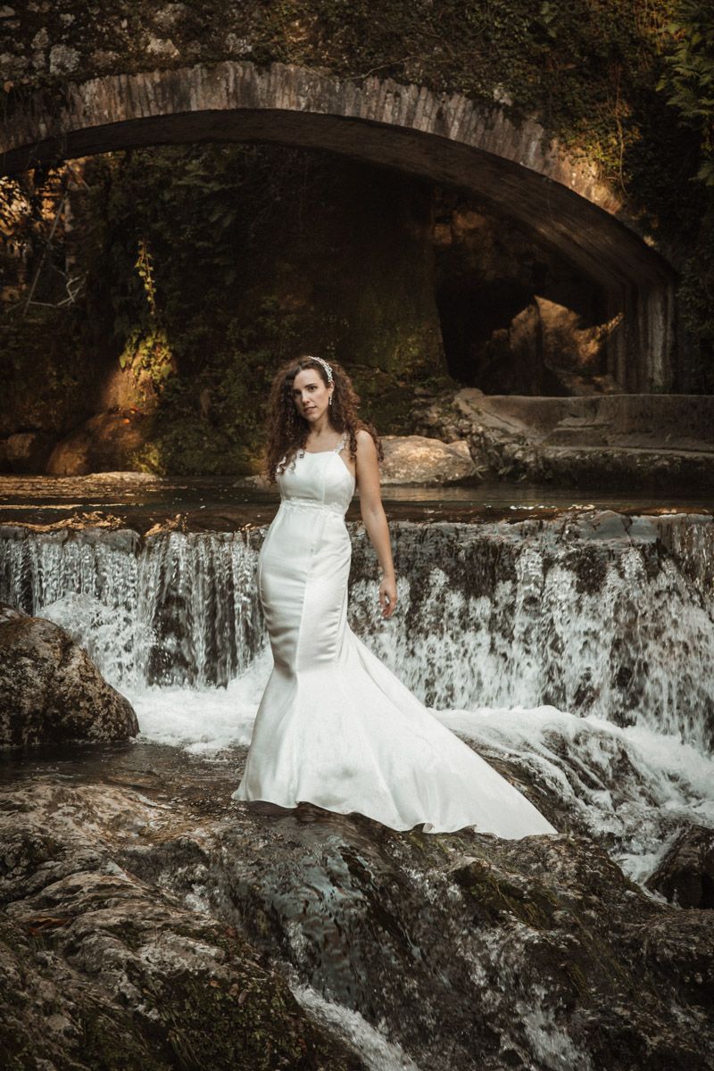 A person in a white wedding dress poses gracefully on rocks near a cascading waterfall in a romantic outdoor setting.