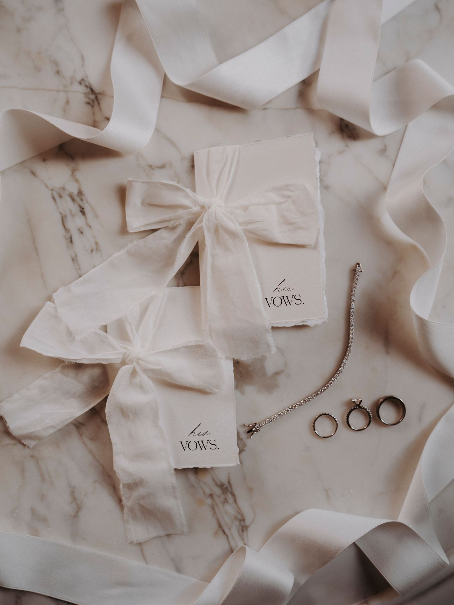 Elegant wedding flat lay with vow books tied in silk ribbons, gold rings, and bracelet on marble surface.