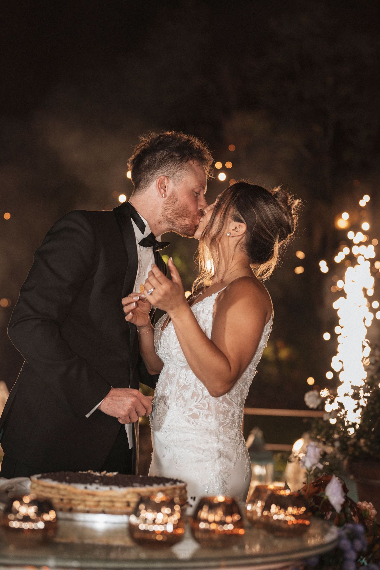 Wedding couple sharing dessert at reception with sparklers glowing in background.