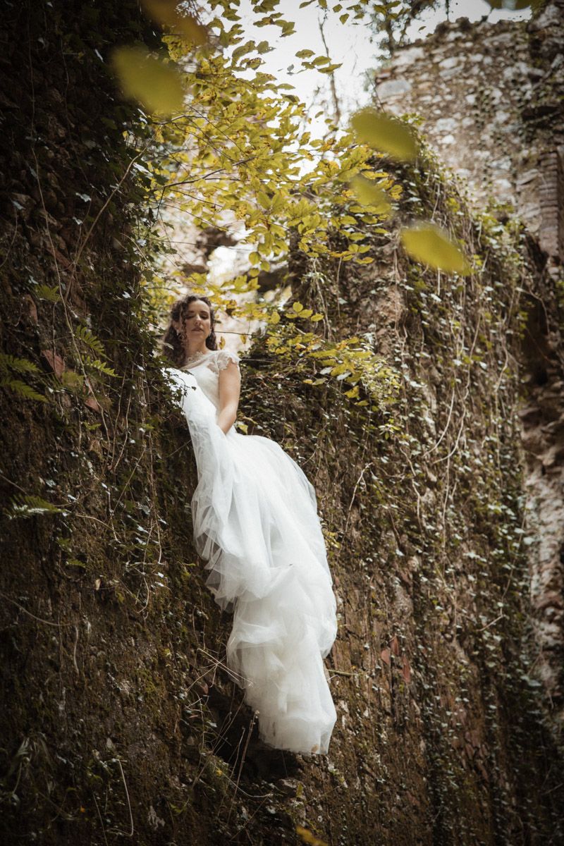 A person in a flowing white wedding dress poses dramatically against a rustic stone wall surrounded by autumn foliage.