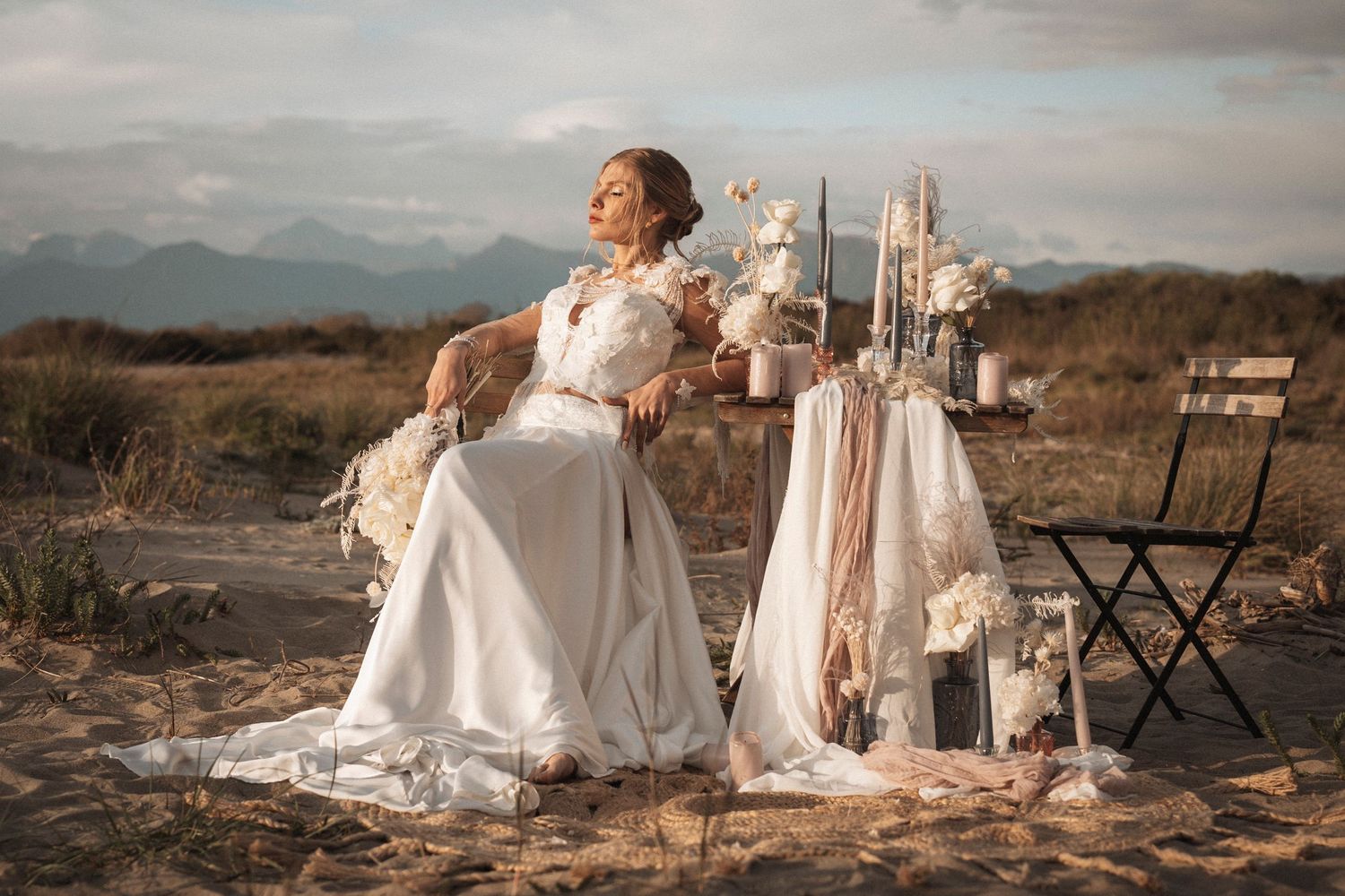 A bride in a flowing white dress poses gracefully in a desert landscape at sunset with vintage decor.
