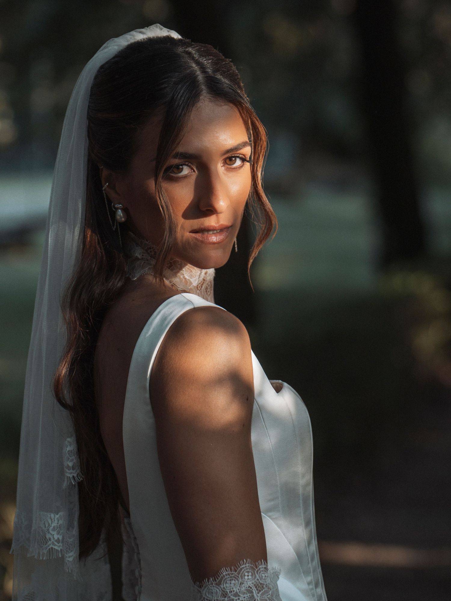 Elegant bride in white gown and veil poses outdoors in golden sunlight, looking over her shoulder.