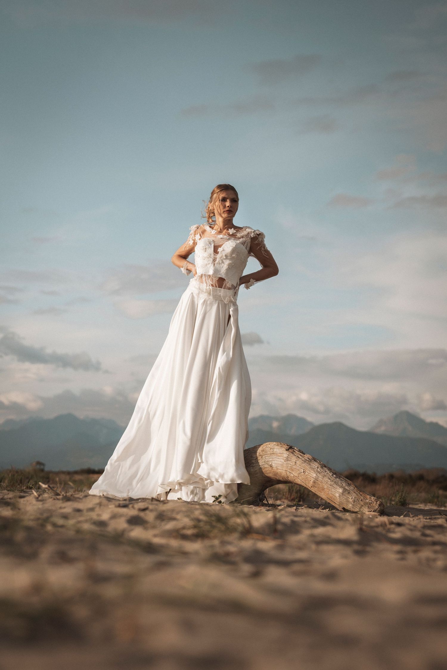 A white wedding dress flows elegantly against a desert landscape with mountains in the background at sunset.
