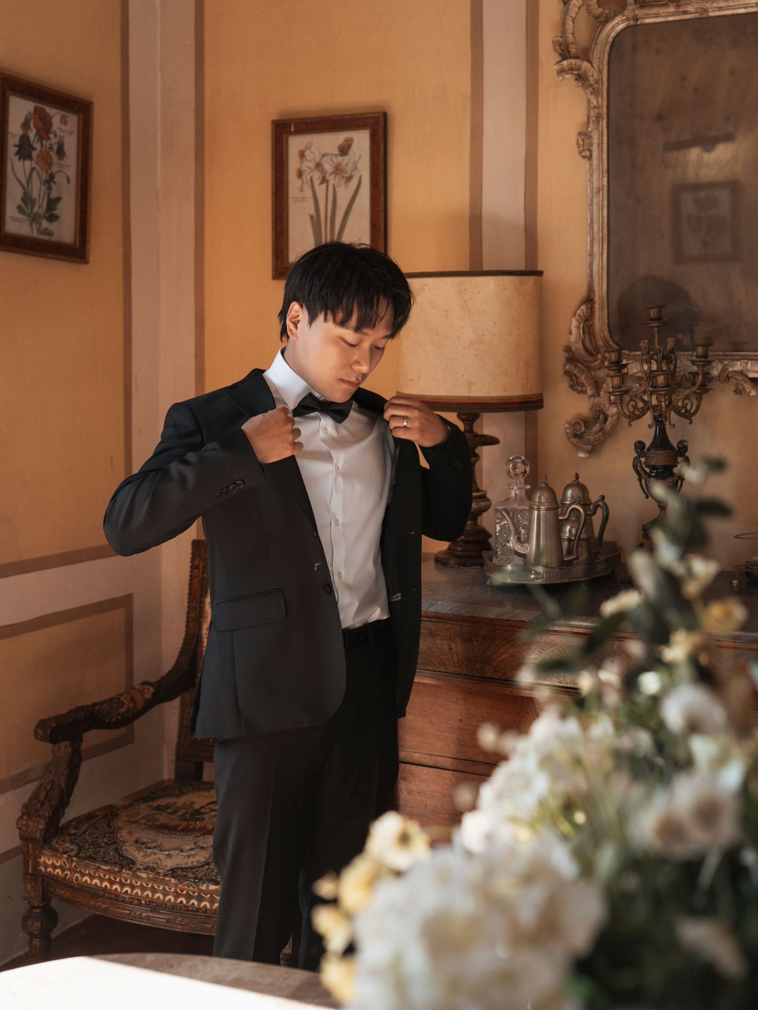 A groom in a black tuxedo adjusts his bow tie in an elegant, warmly lit room with floral decor.