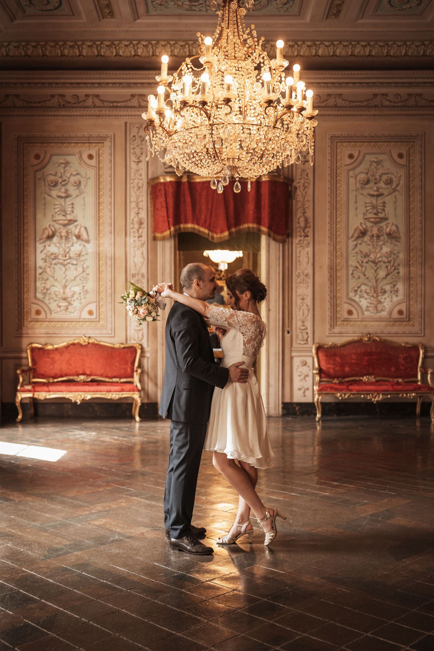 Couple dancing in an ornate ballroom with crystal chandeliers and vintage furniture.