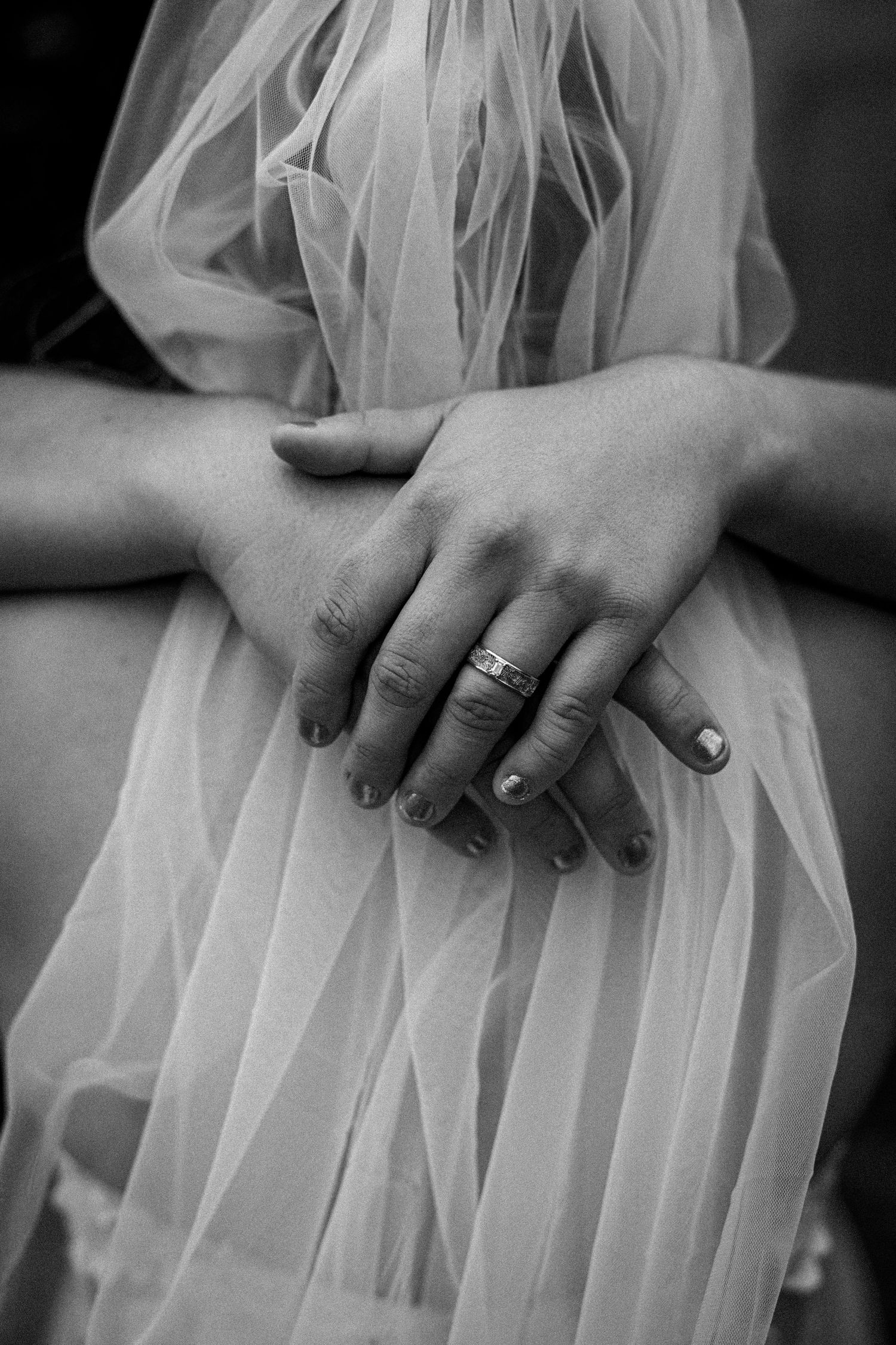 Close-up black and white photo of hands with wedding ring against flowing white tulle fabric.