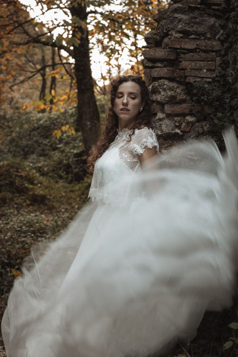 A person in a white lace wedding dress poses dramatically in a misty forest setting during autumn.
