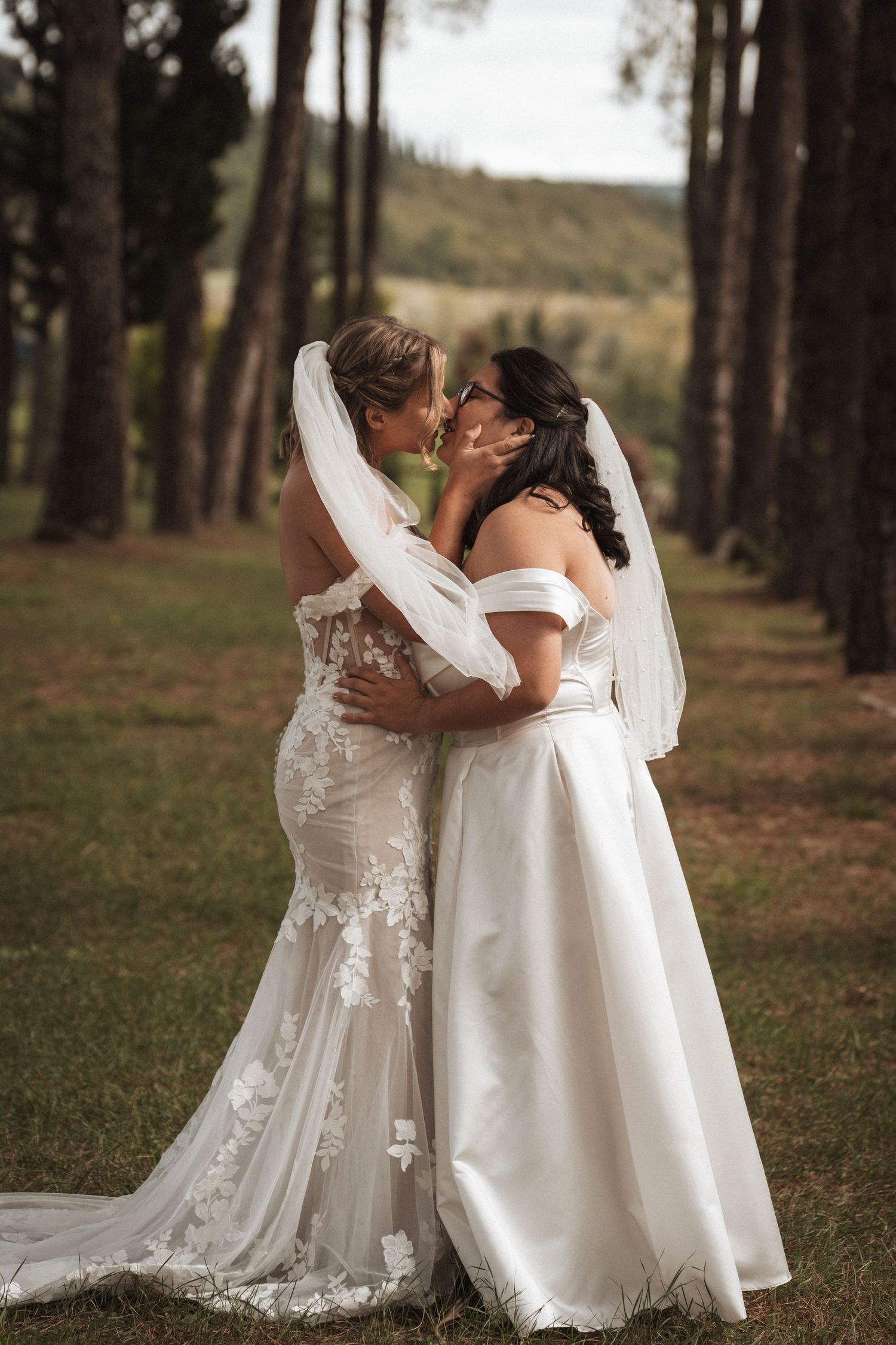 Two brides embracing in romantic forest setting wearing white wedding gowns.
