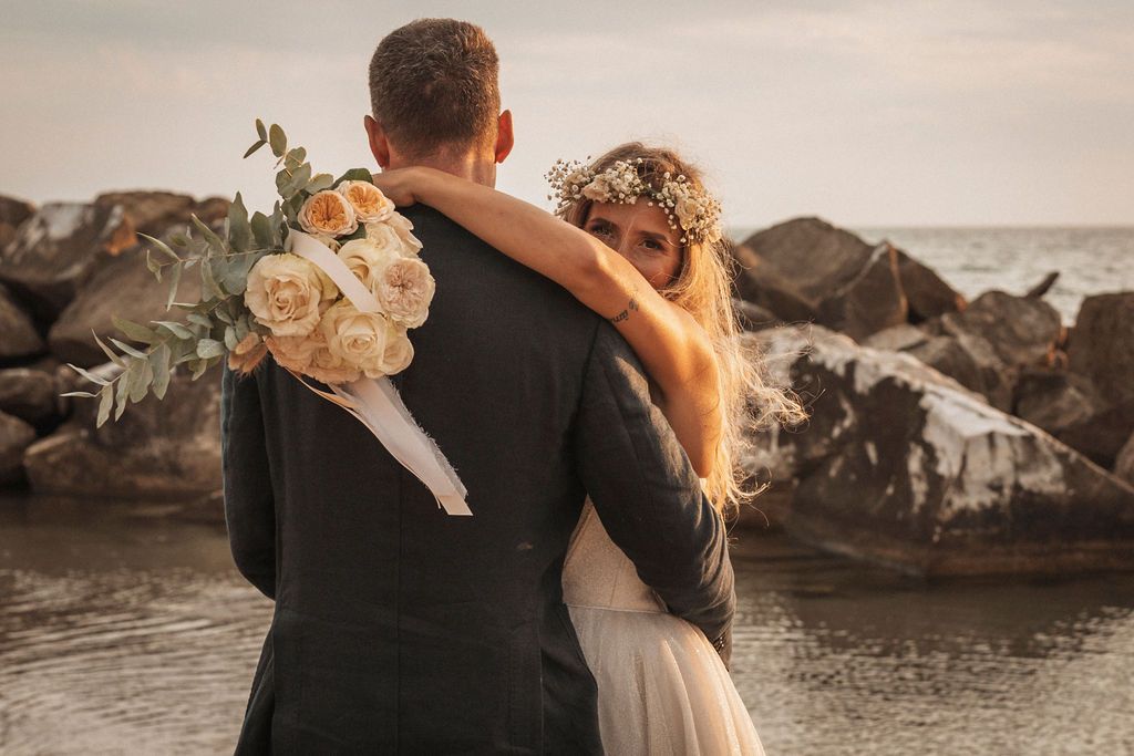 A couple embraces on a rocky beach at sunset with the ocean in the background.