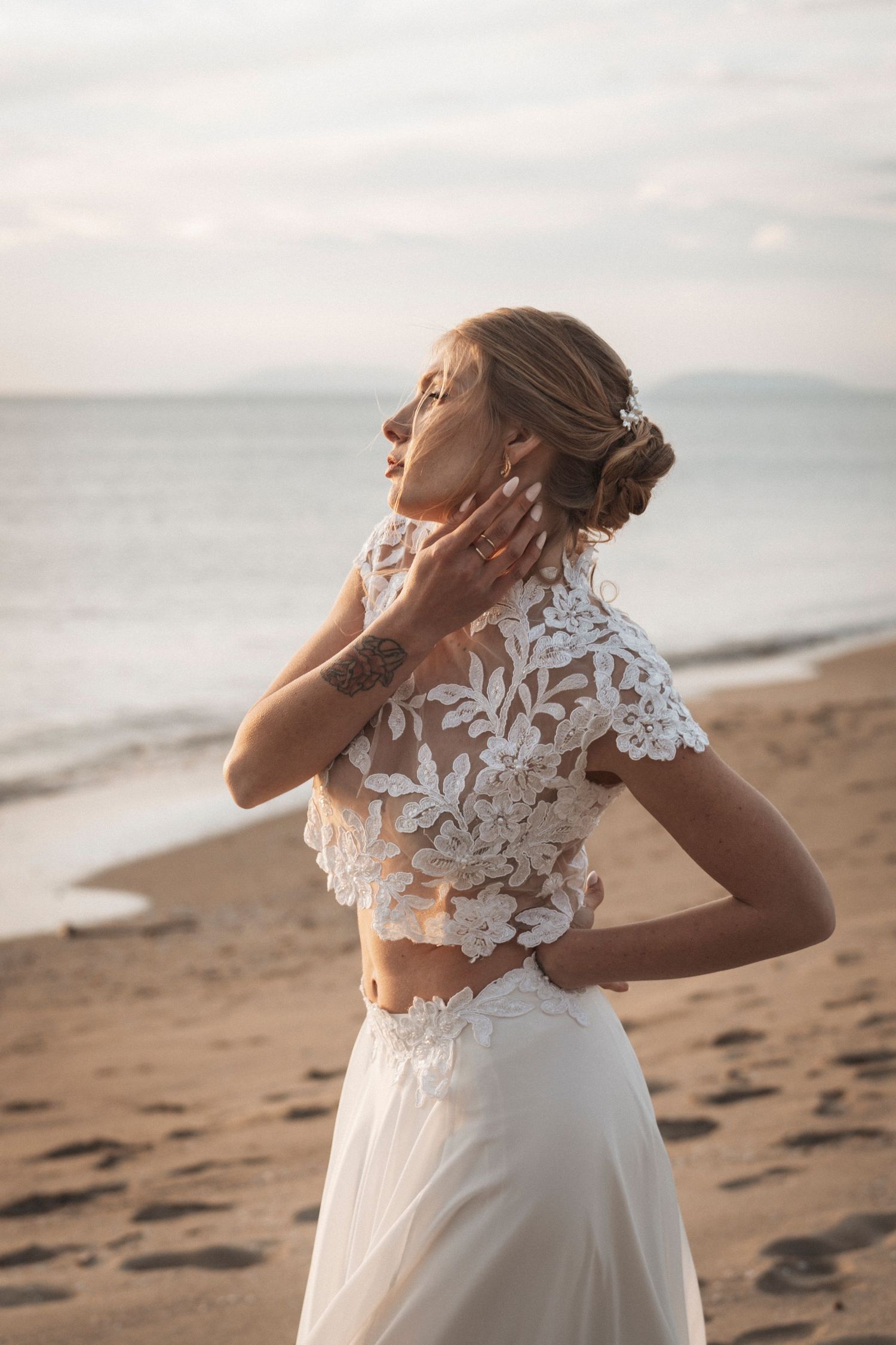 A bride in a white lace dress twirls joyfully on a sandy beach at sunset.