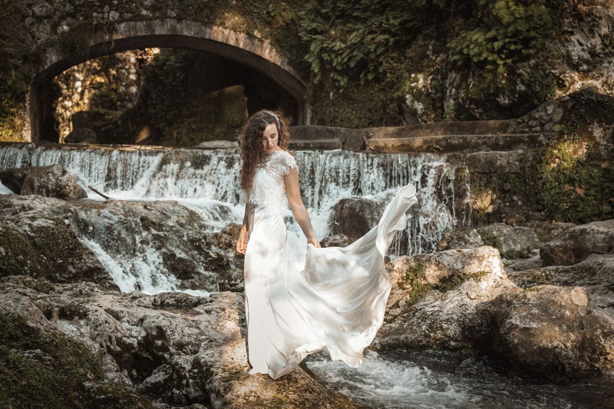 A flowing white wedding dress billows in the breeze against a scenic waterfall backdrop in a natural forest setting.