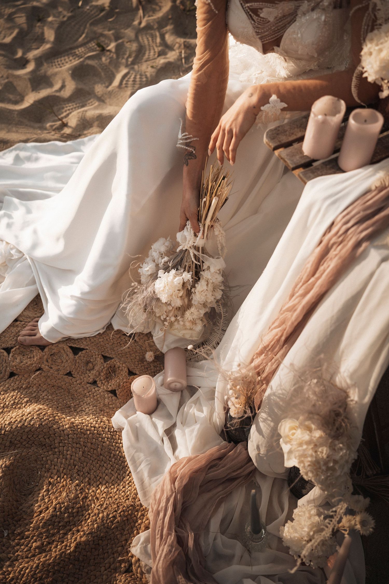 Artistic still life of wedding dress details with dried flowers and candles arranged on sandy beach texture.