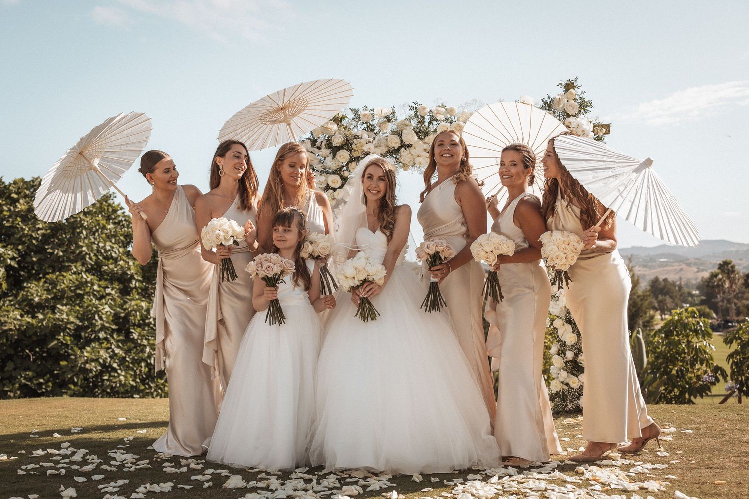 Bridesmaids in light colored dresses pose with white bouquets against a scenic mountain backdrop.