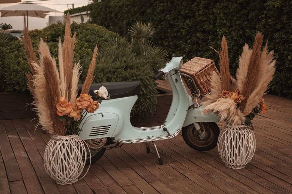 Vintage mint-colored Vespa scooter decorated with pampas grass and woven lanterns on a wooden deck.