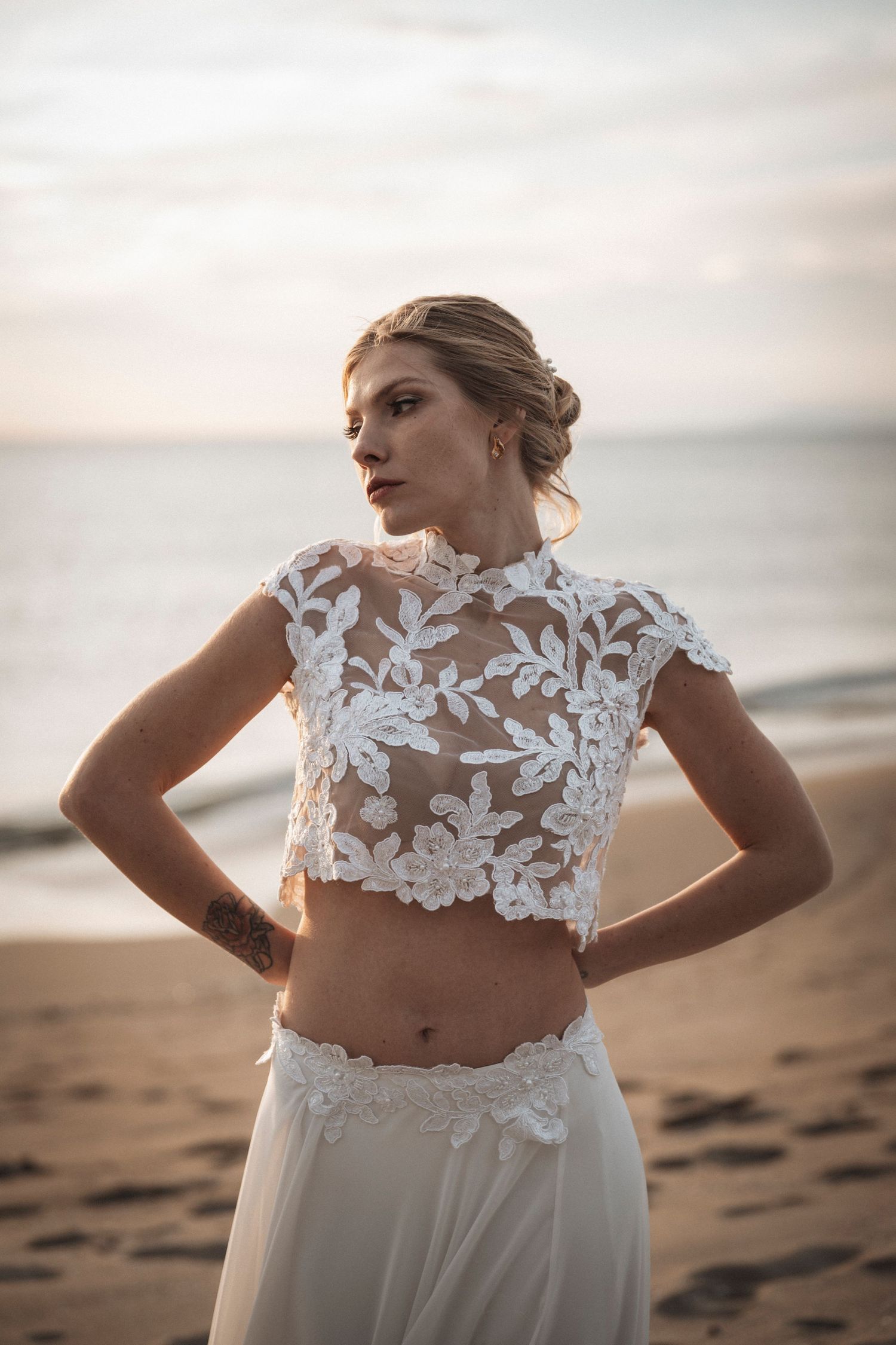 Delicate white lace crop top wedding dress with floral appliques photographed on a sandy beach at dusk.