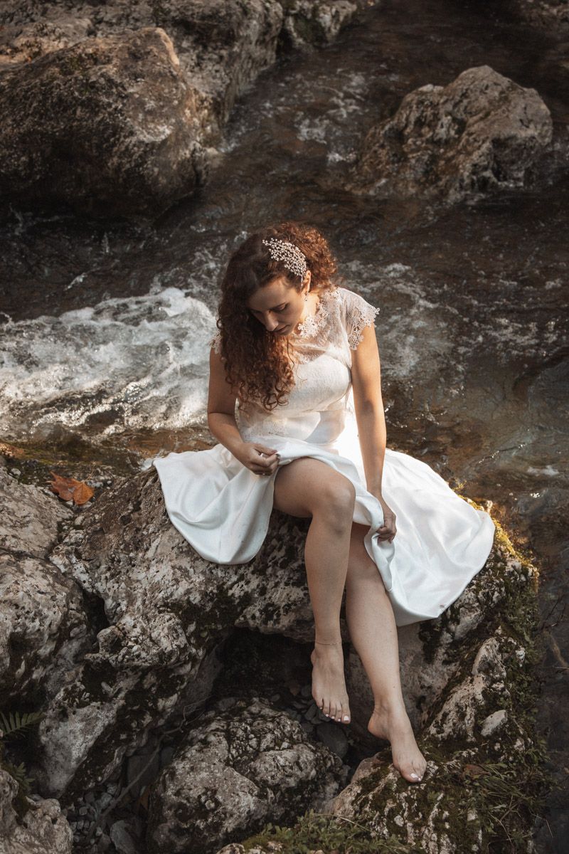 A person in a white dress sits barefoot on coastal rocks with crashing waves in the background.