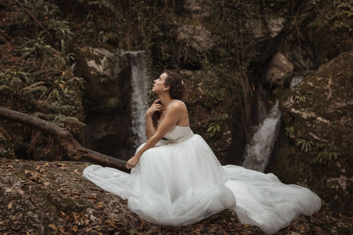 A flowing white wedding dress cascades over mossy rocks near a waterfall in a mystical forest setting.