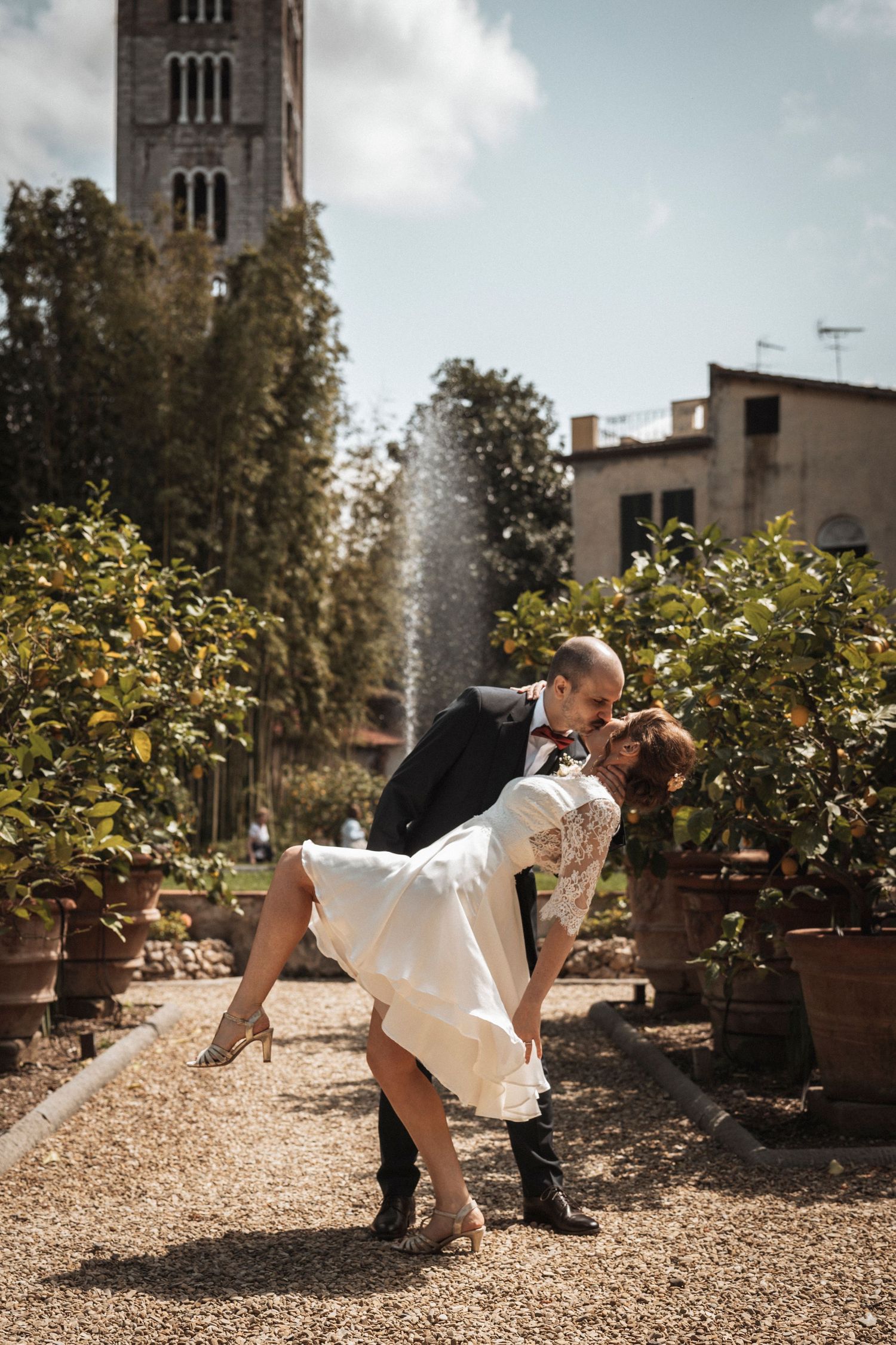 Wedding couple posing playfully in a sunny garden with fountain and potted plants.