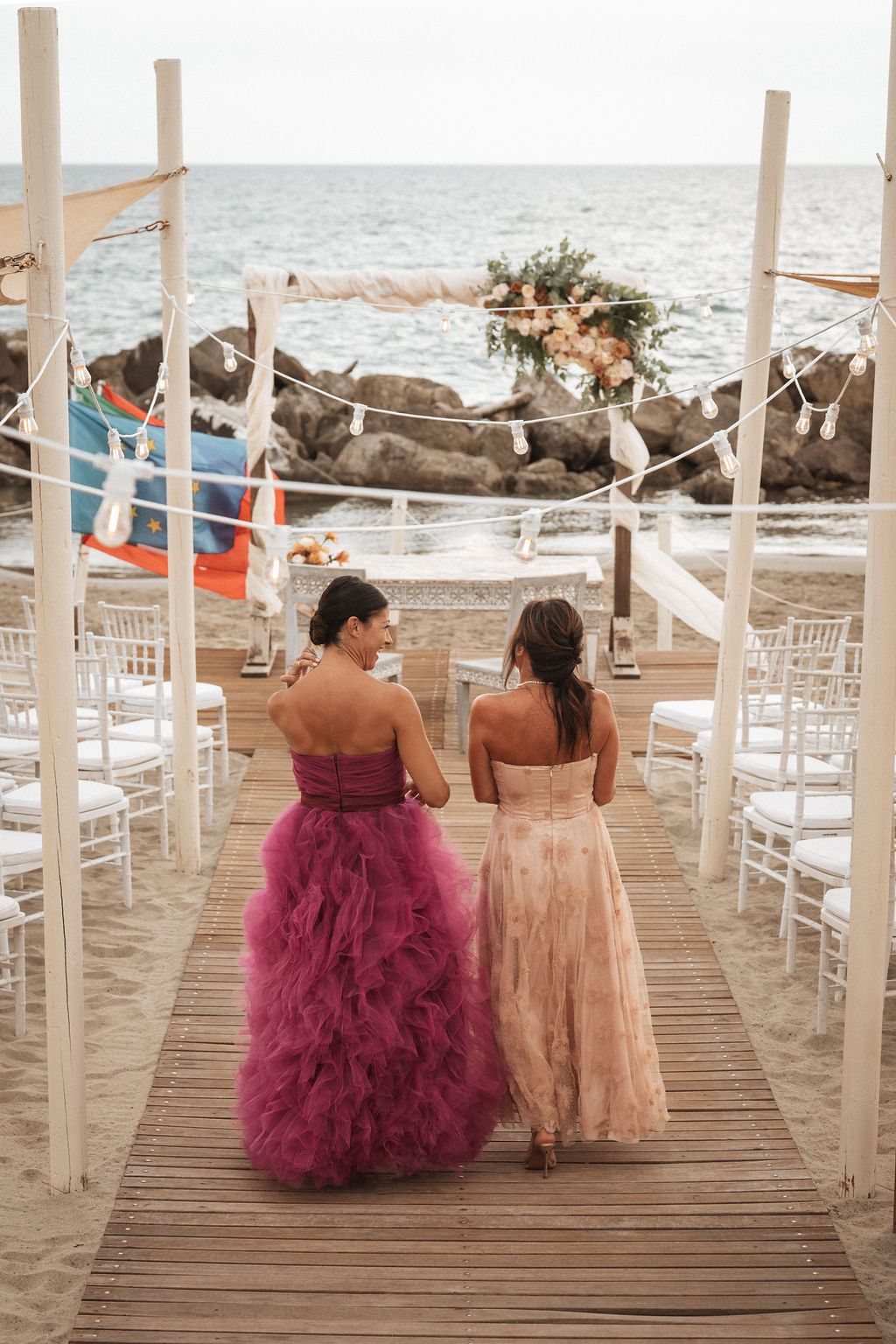 Beach wedding setup with white chairs and two guests in pink and peach formal dresses viewing the ocean.